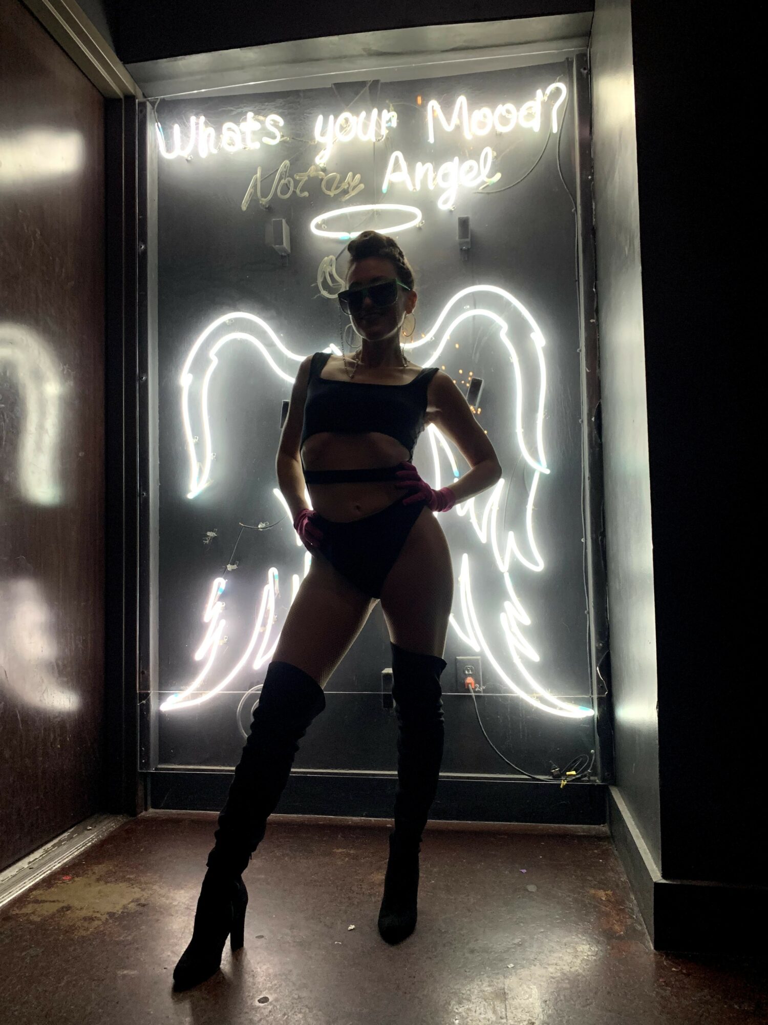 Person standing in front of neon angel wings display, wearing sunglasses, a striped top, shorts, and thigh-high boots, with a sign above reading 'What's your Mood?'.