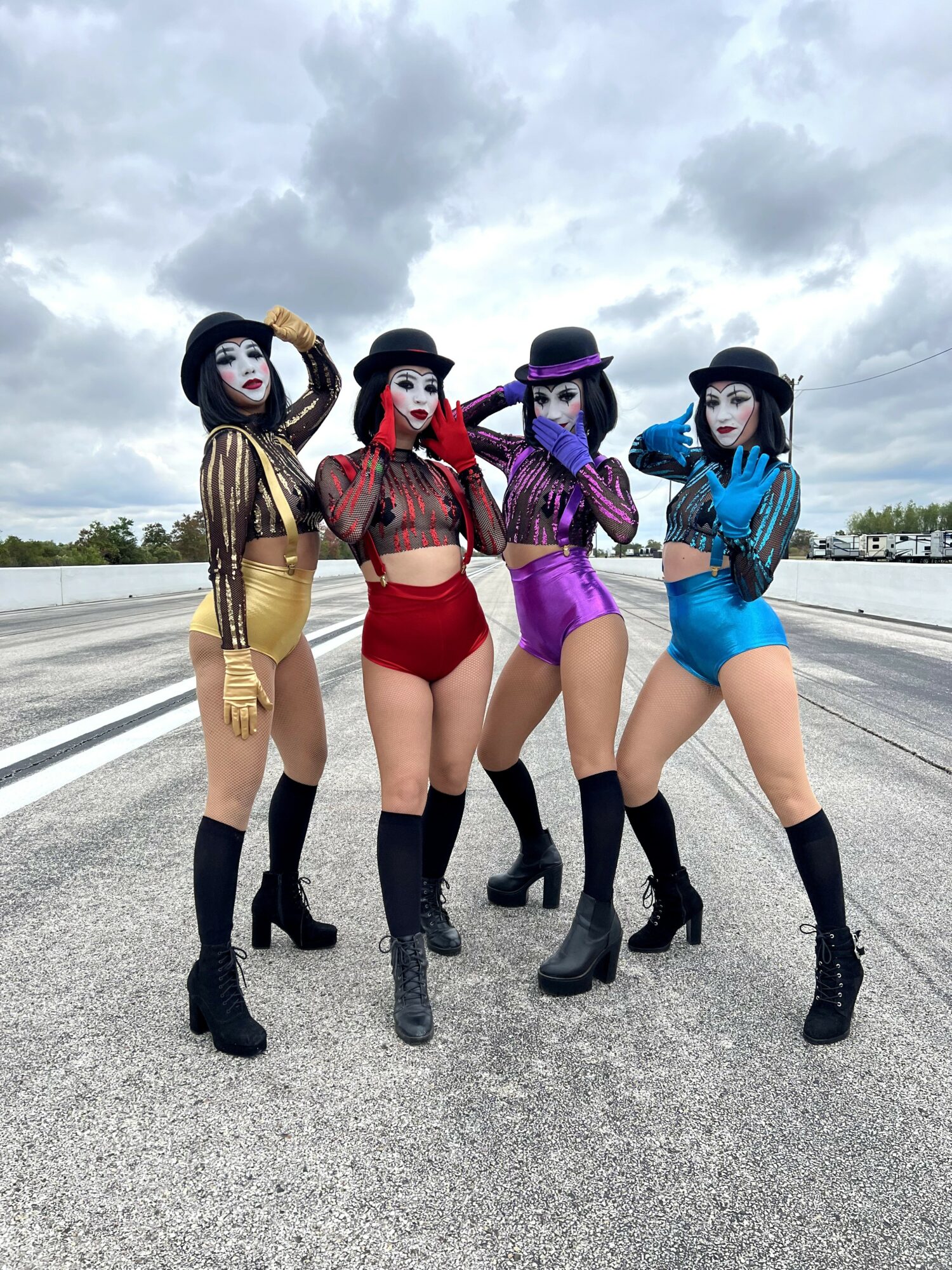 Four women in colorful costumes and hats standing on an empty road with cloudy sky background.