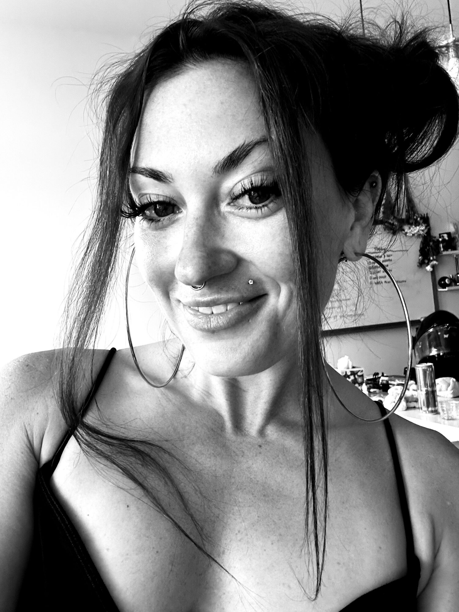 Young woman with dark hair styled in buns, smiling, wearing a tank top, in a room with various objects in the background.