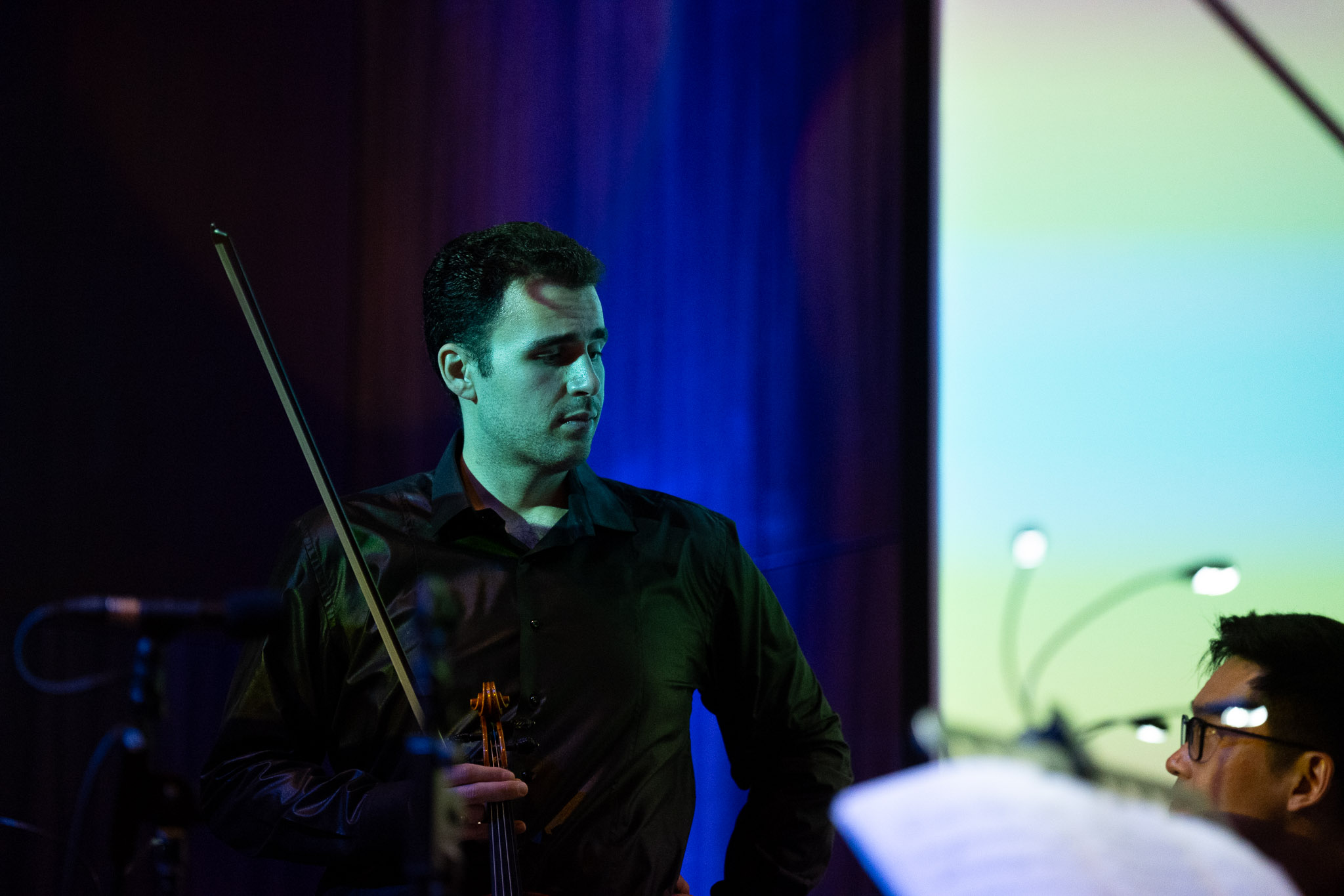Man holding a violin bow, looking to the side, with a colorful background and a person wearing glasses in the foreground.