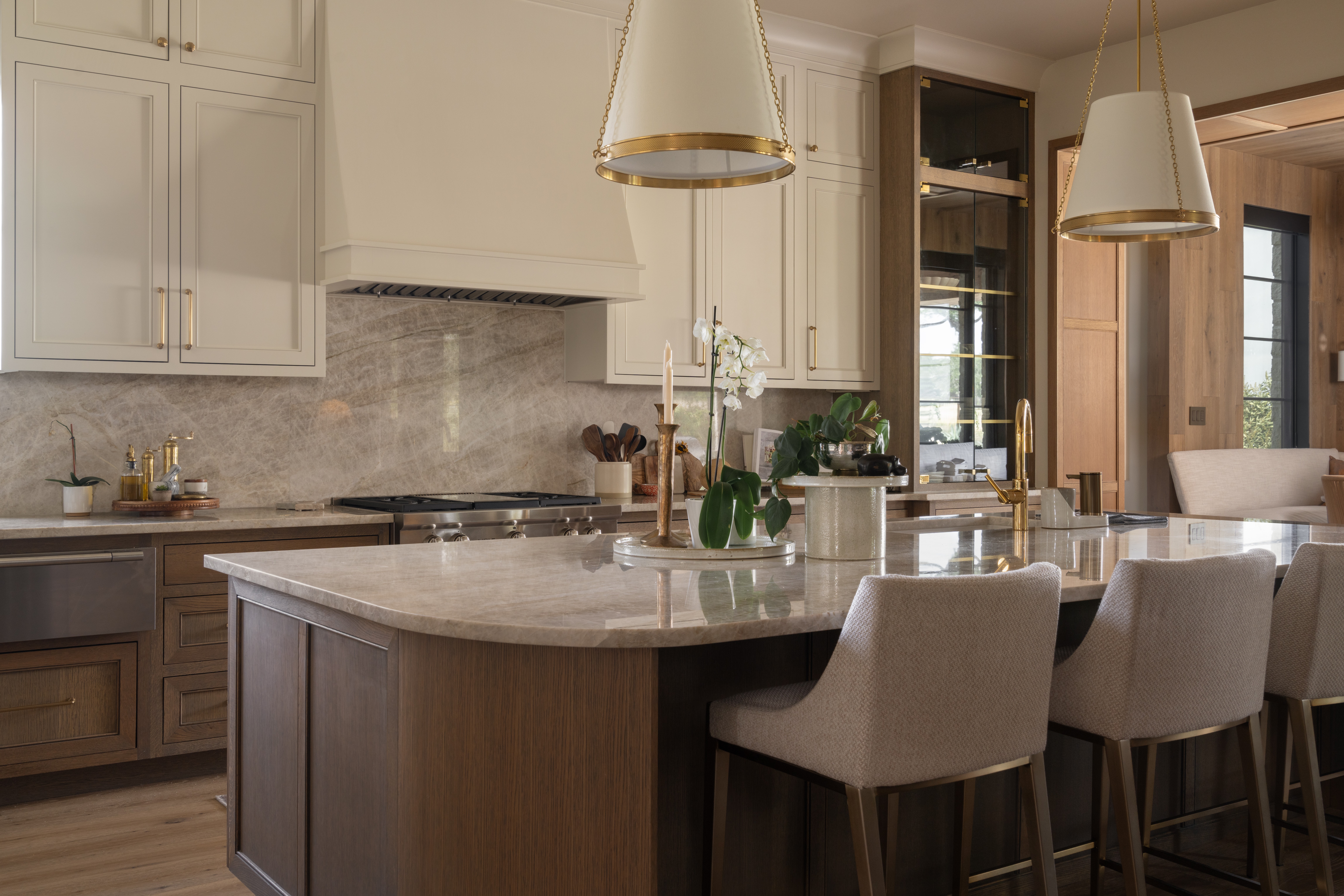 Modern kitchen with white cabinets, a large island with a marble countertop, and three beige barstools, illuminated by pendant lights.