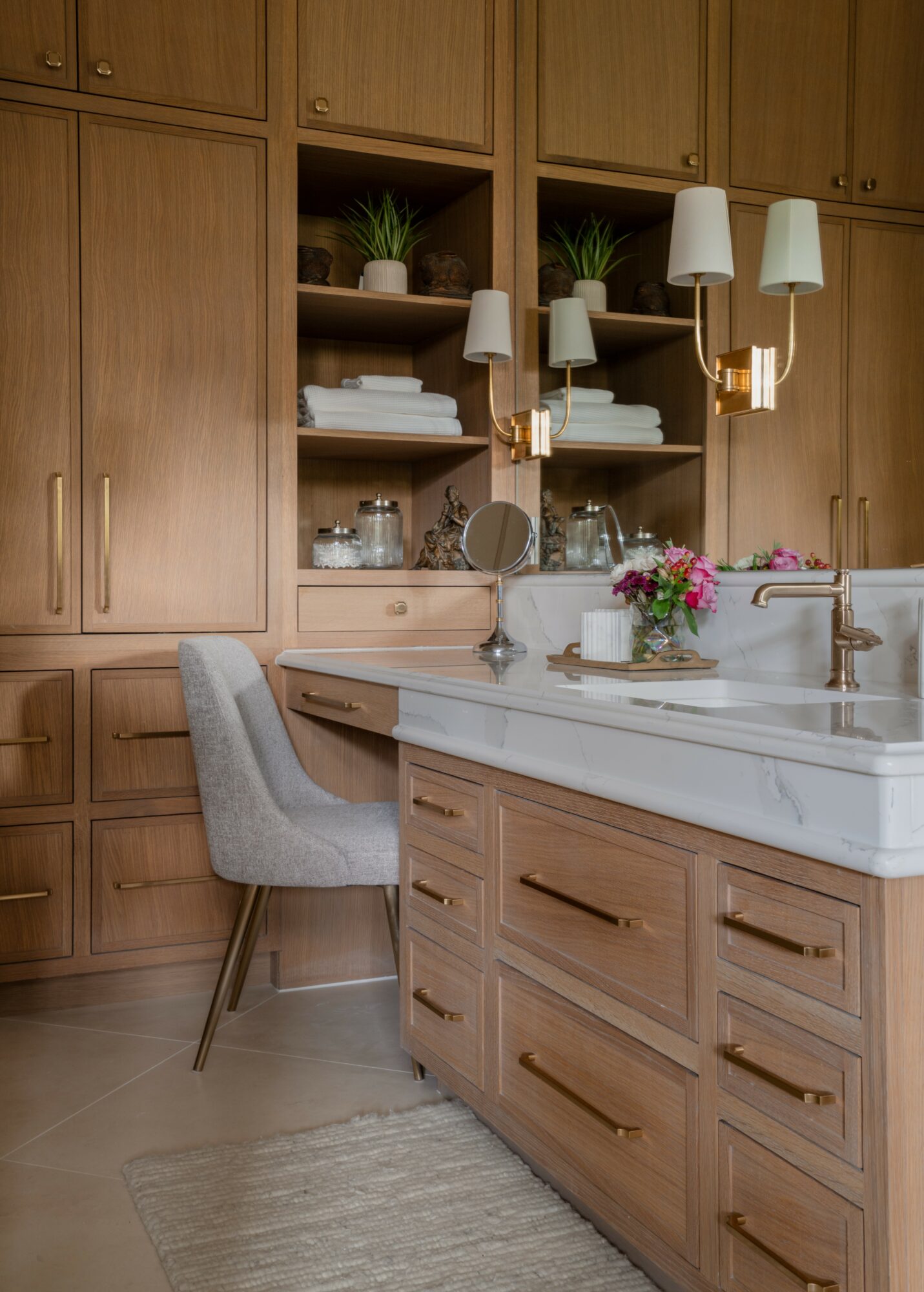 Wooden kitchen cabinets with open shelves, a white marble countertop, a gray chair, and a vase with pink flowers.