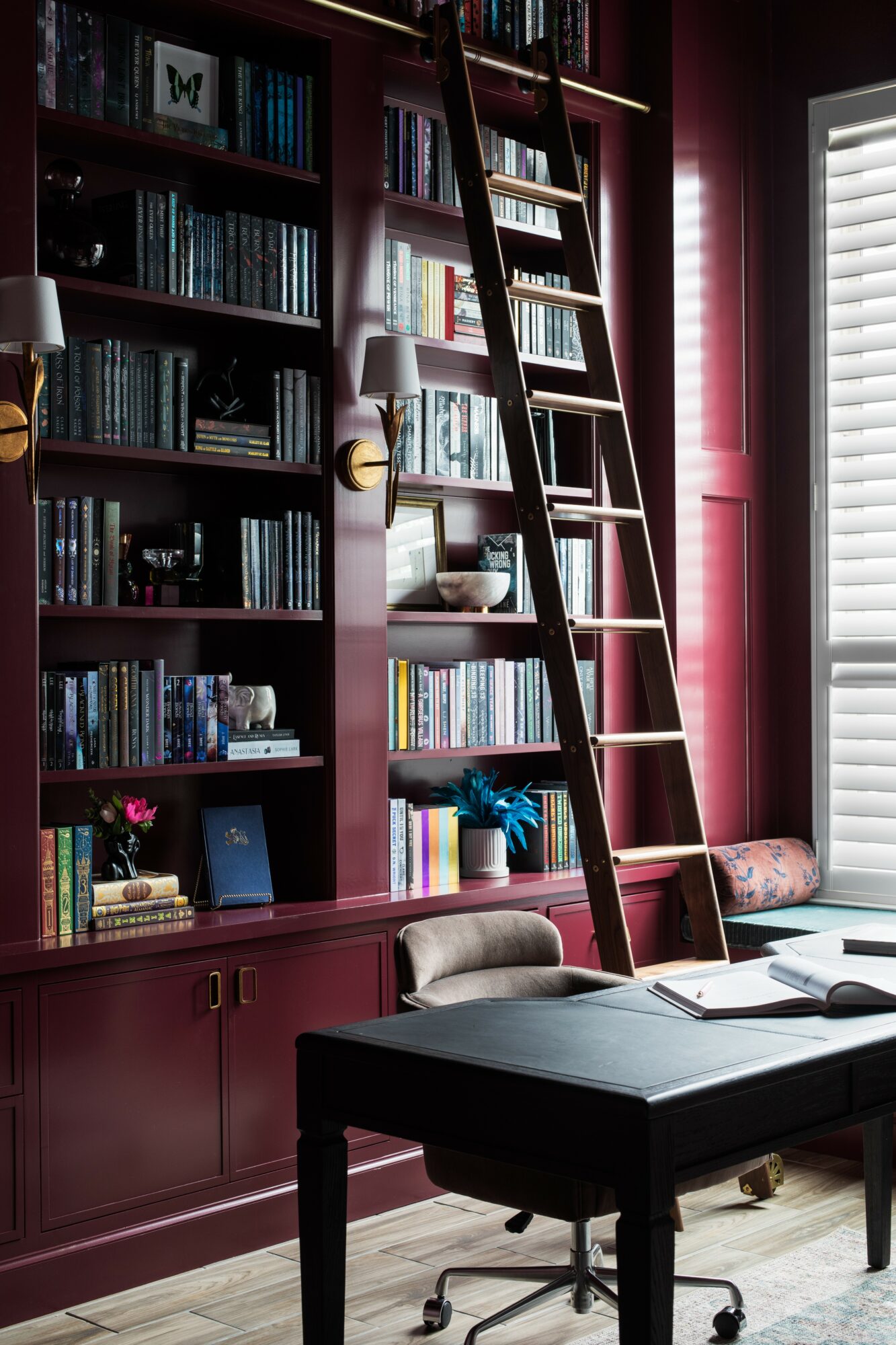 Bookshelf with books, ladder, window with blinds, and a black desk with a chair in a room with red walls.