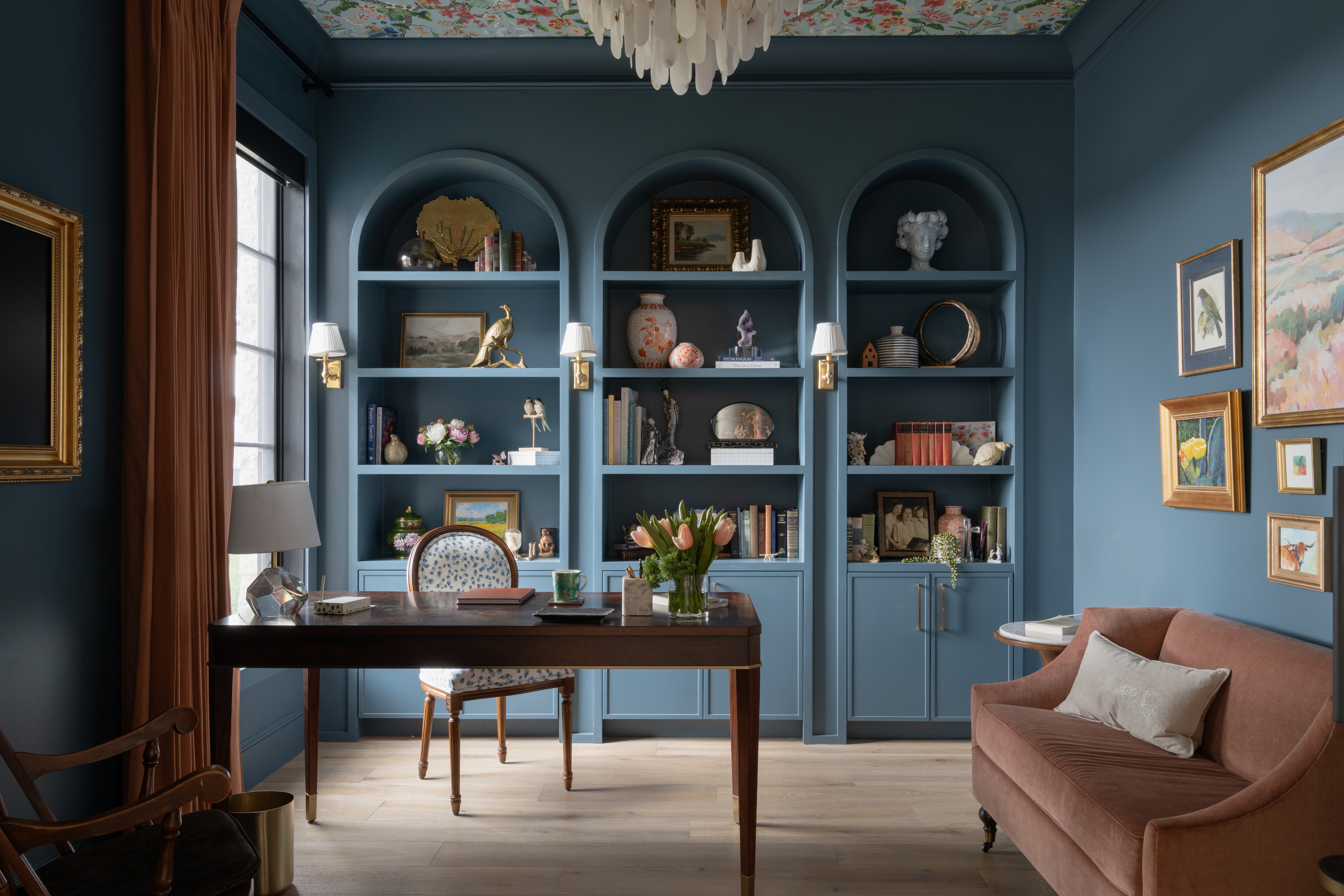 Elegant room with blue built-in bookshelves, a wooden desk, a pink sofa, and framed artwork on the wall.