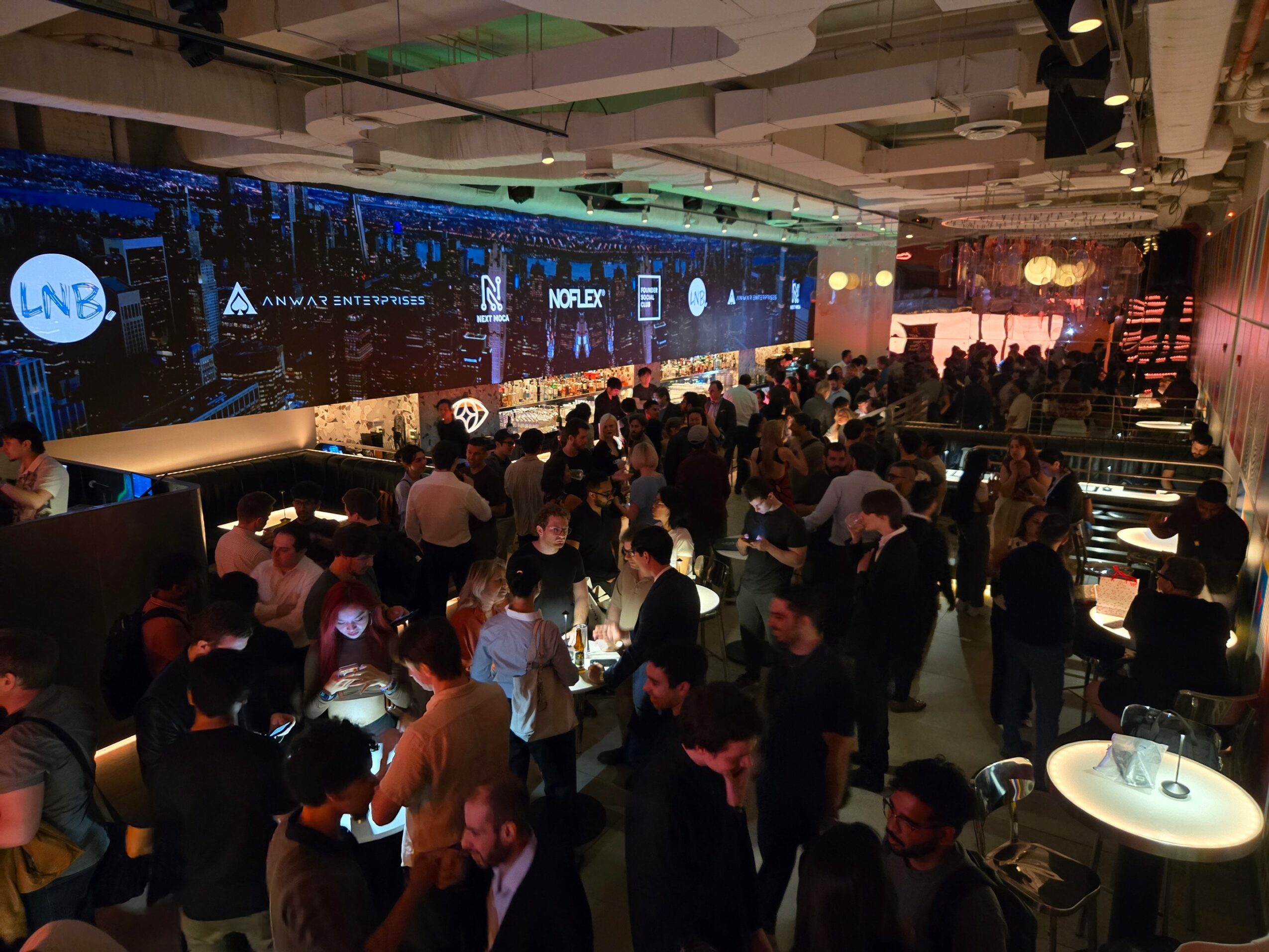 Crowd of people at an indoor event with a large stage and screens, some standing and some seated, with dim lighting.