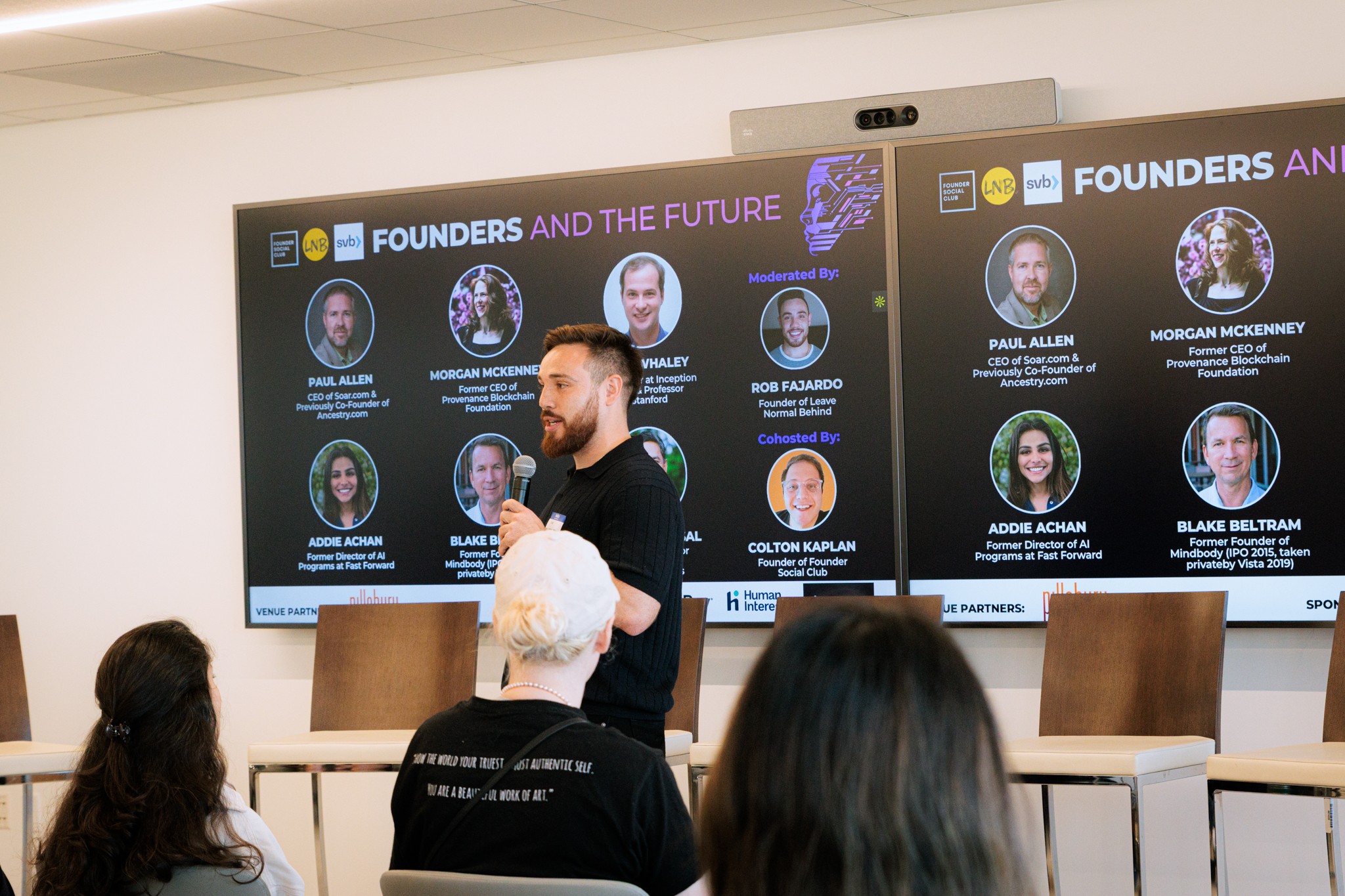 A man speaking in front of a large screen displaying a panel of speakers and event information, with an audience listening.