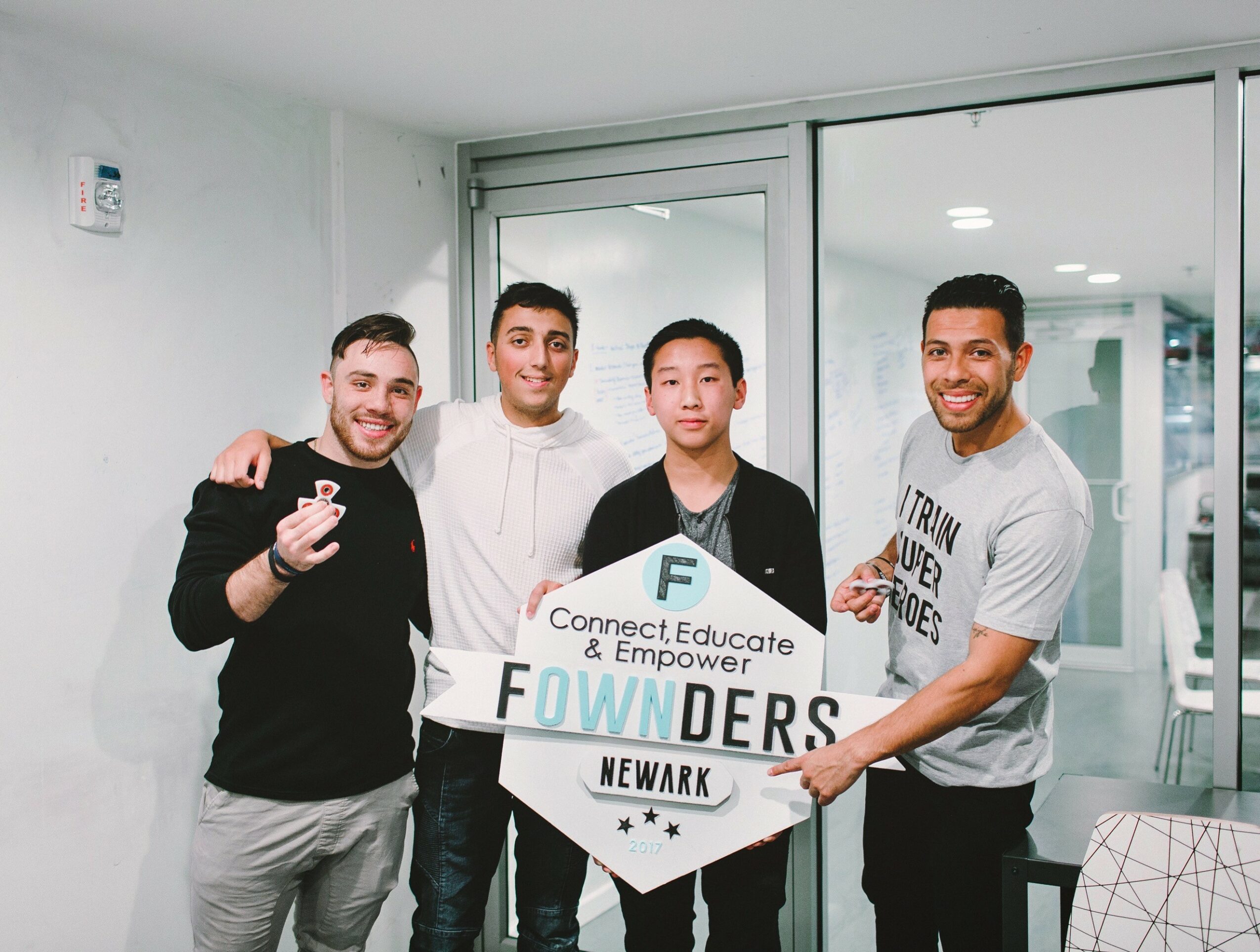 Four people holding a sign that reads 'Connect, Educate, Empower, FOWNDERS, NEWARK' in a room with glass doors.