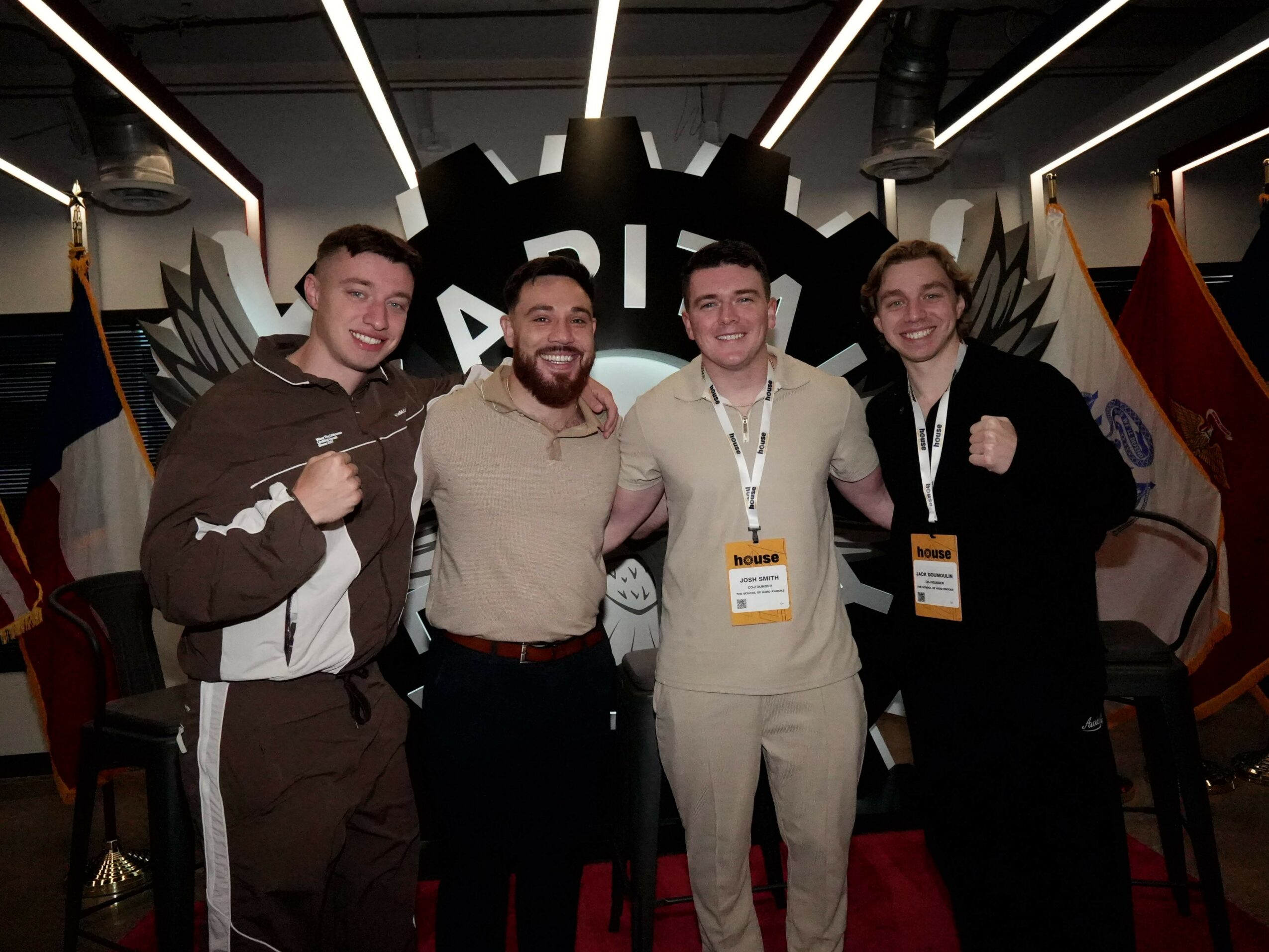 Four men standing together in front of a gear-shaped backdrop with flags on either side, smiling and posing for the photo.