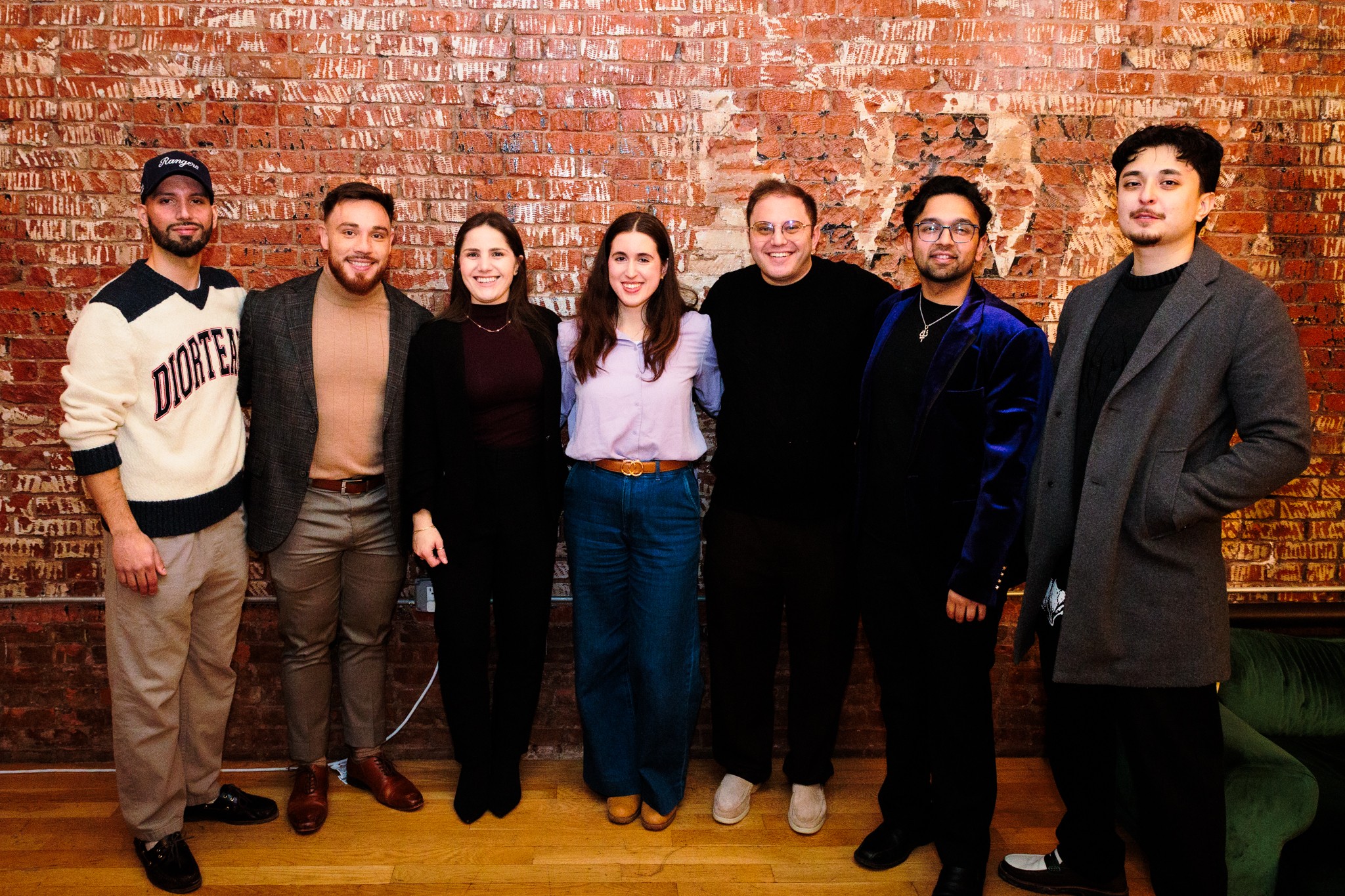 Group of seven people standing in front of a brick wall, smiling, dressed casually, indoors.