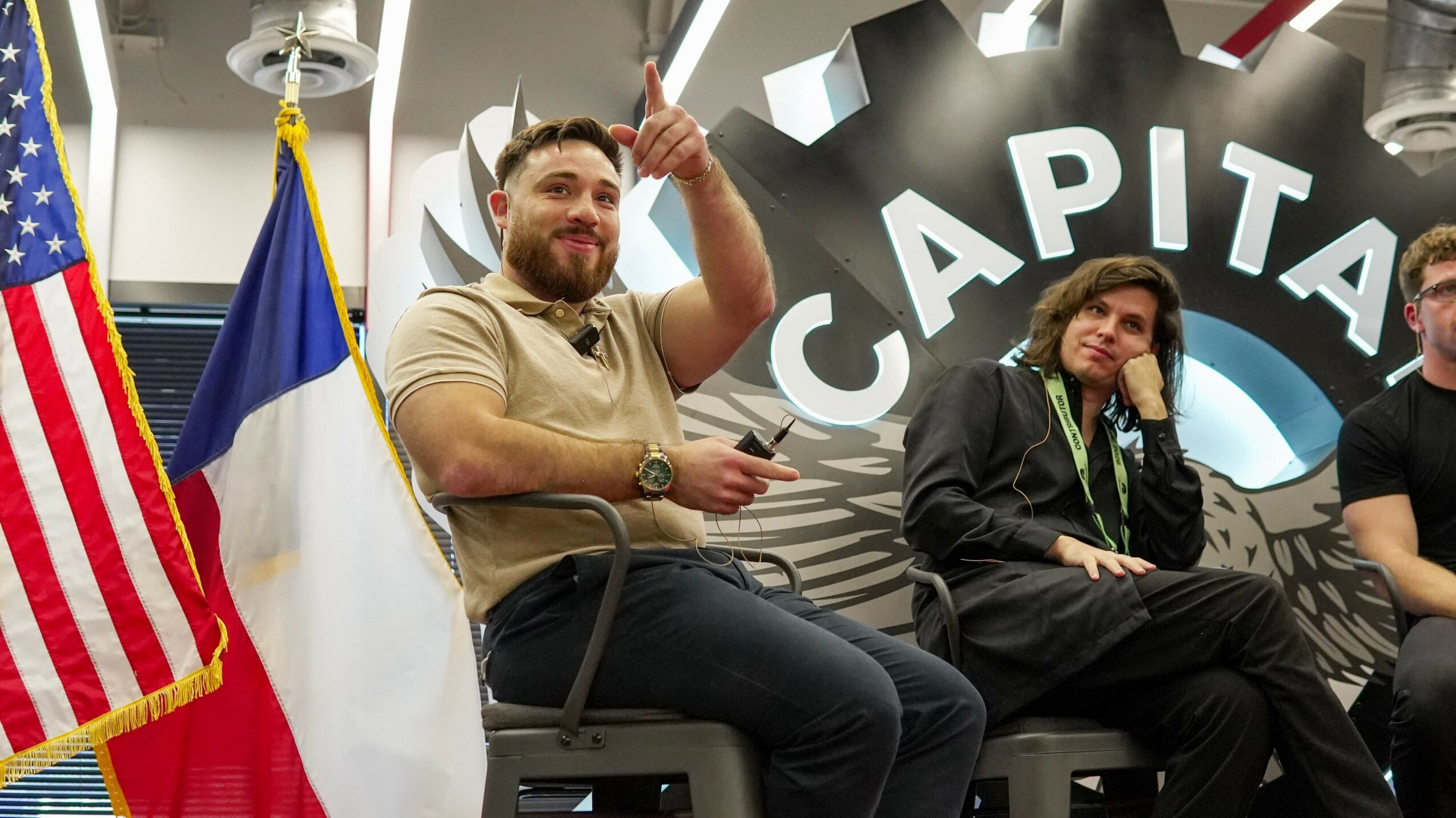 Two people sitting on a panel, one gesturing with a raised finger, with flags and a large 'CAPITOL' sign behind them.