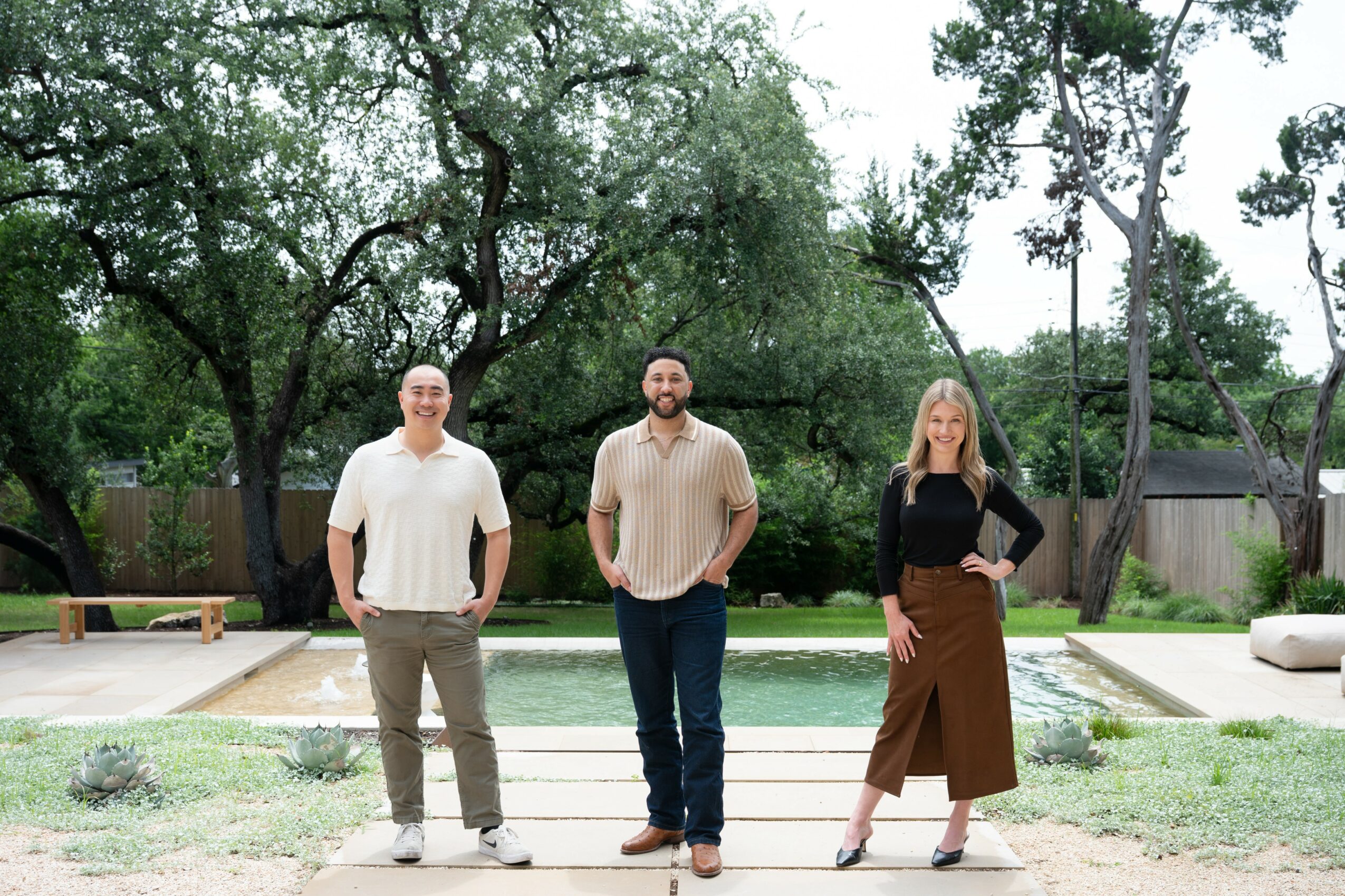 Three people standing outdoors in front of a pool, trees, and a grassy area, smiling and posing for the photo.