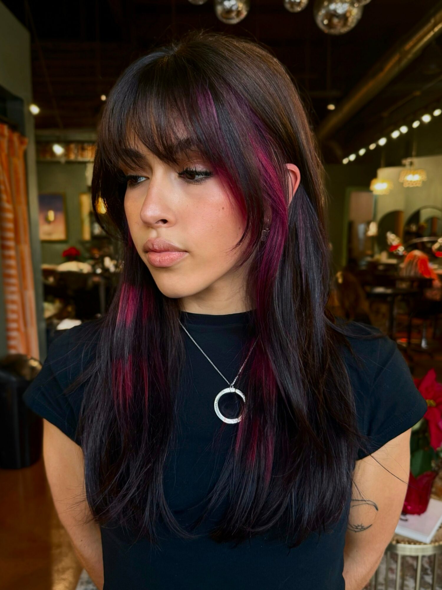 Young woman with long dark hair and purple highlights, wearing a black top and a circular pendant necklace, in a cafe.