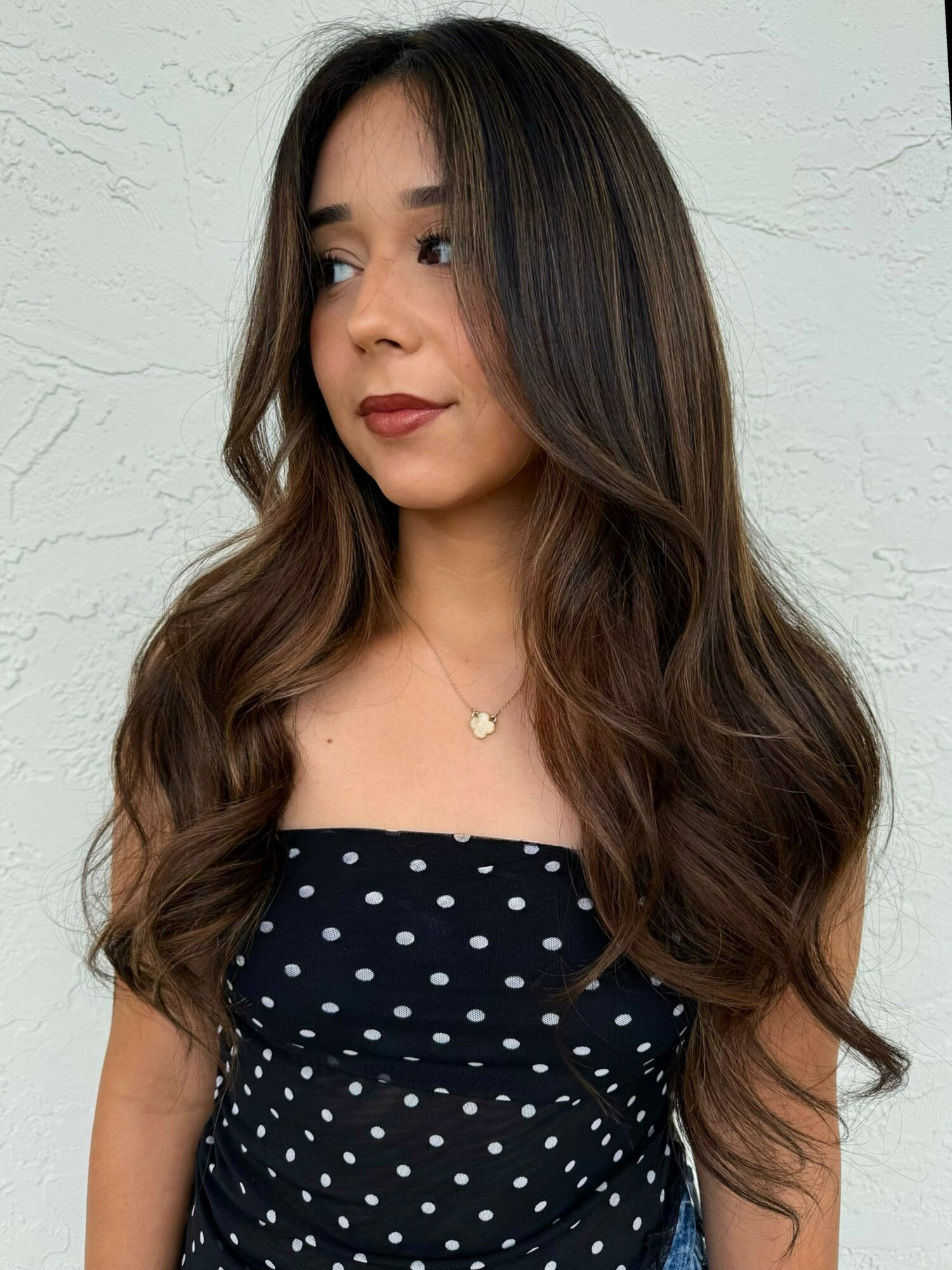 Young woman with long wavy hair wearing a black polka dot top and a necklace, looking to the side.