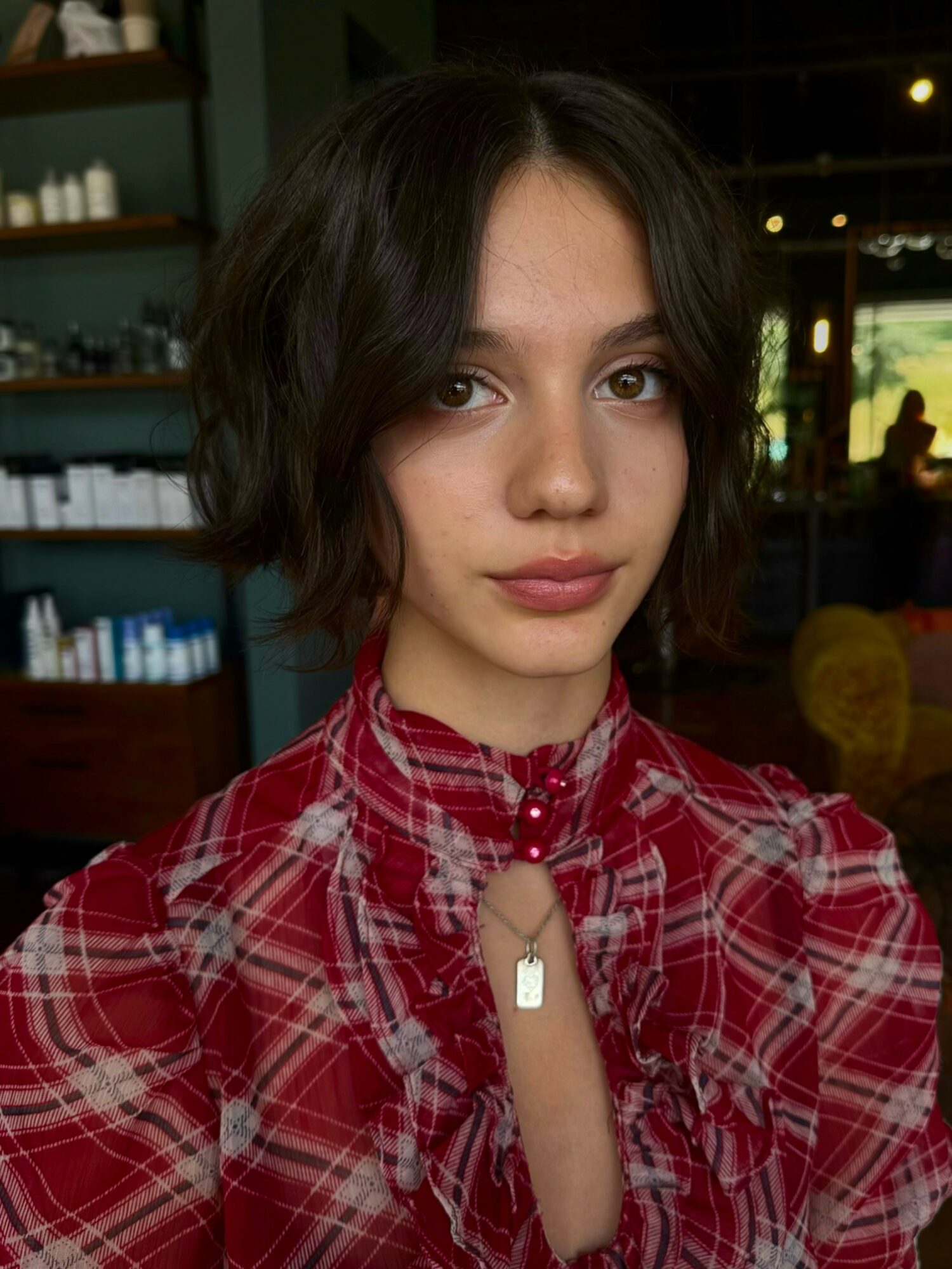 Young woman with short dark hair wearing a red plaid shirt indoors, looking at the camera.