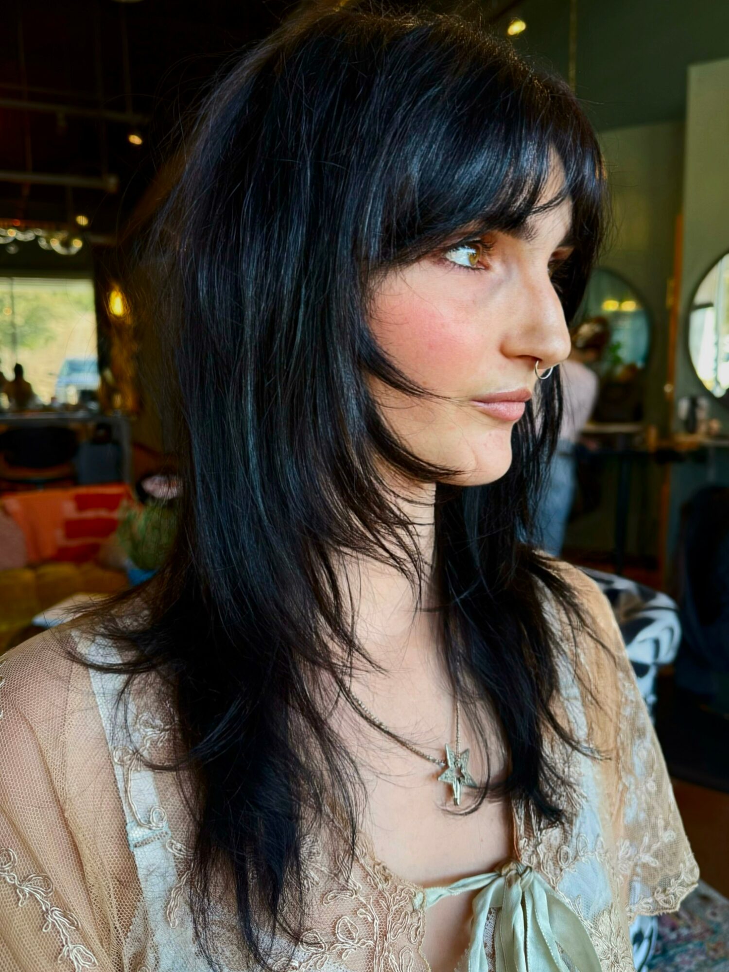 Side profile of a woman with long, dark hair and light skin, wearing a beige top and a star-shaped necklace, indoors.