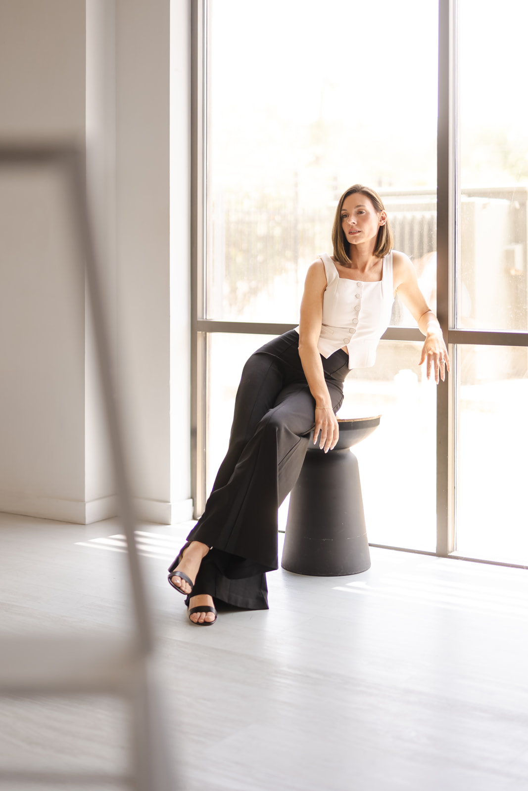 Woman sitting on a stool near large window, sunlight streaming in, wearing a white top and black pants.