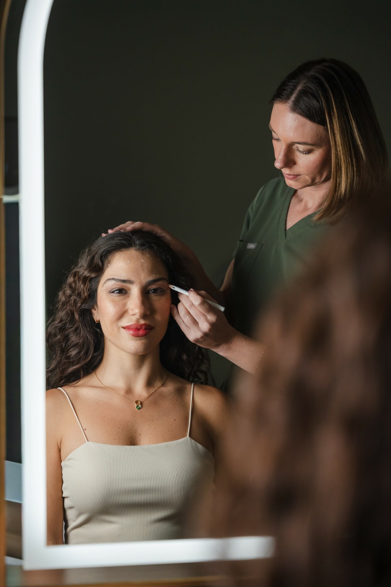 Woman with curly hair and red lipstick sits in front of mirror, makeup artist applies makeup, dark background, woman wears white top