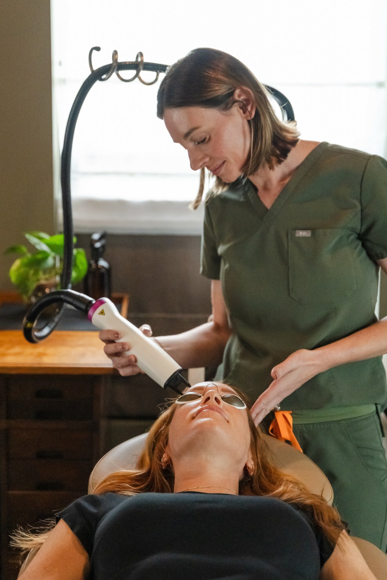 A woman lying on a treatment bed receiving a facial, with another woman in medical scrubs applying a device to her face.