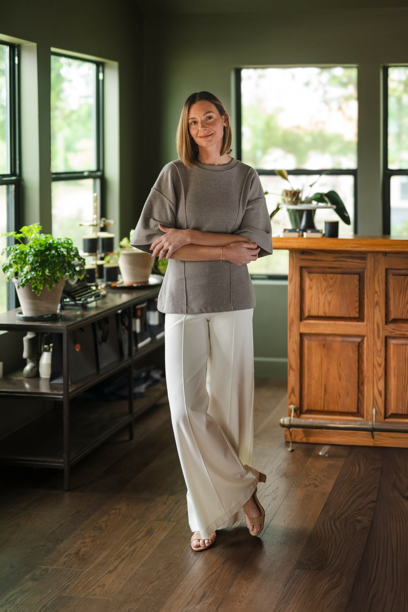 Woman standing in a room with large windows, wearing a gray top and white pants, with plants and furniture nearby.