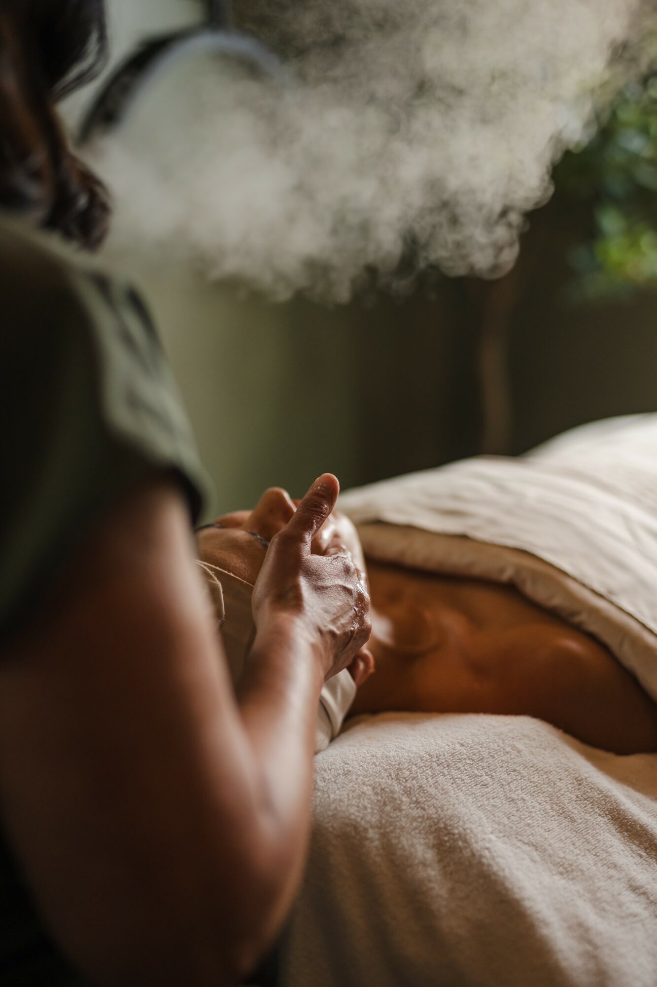 Person holding a glass with vapor or smoke, with a bed and blanket in the background.