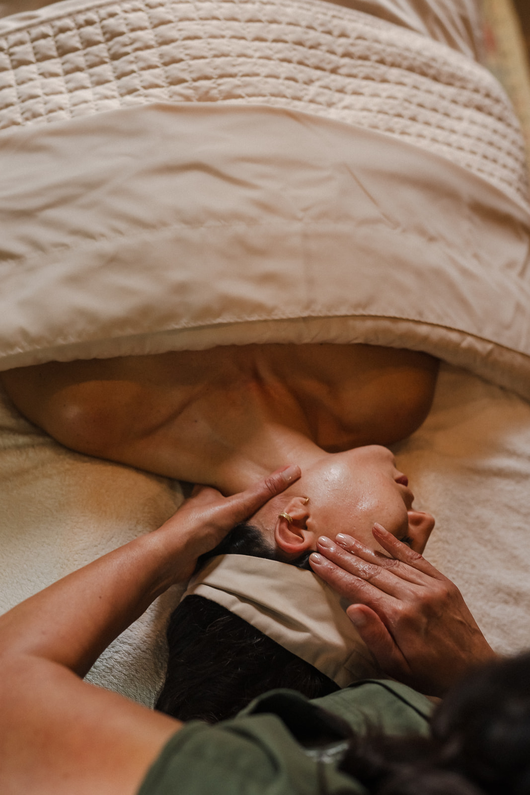 Person lying on bed with head under blanket, hands gently touching face, in a cozy setting.
