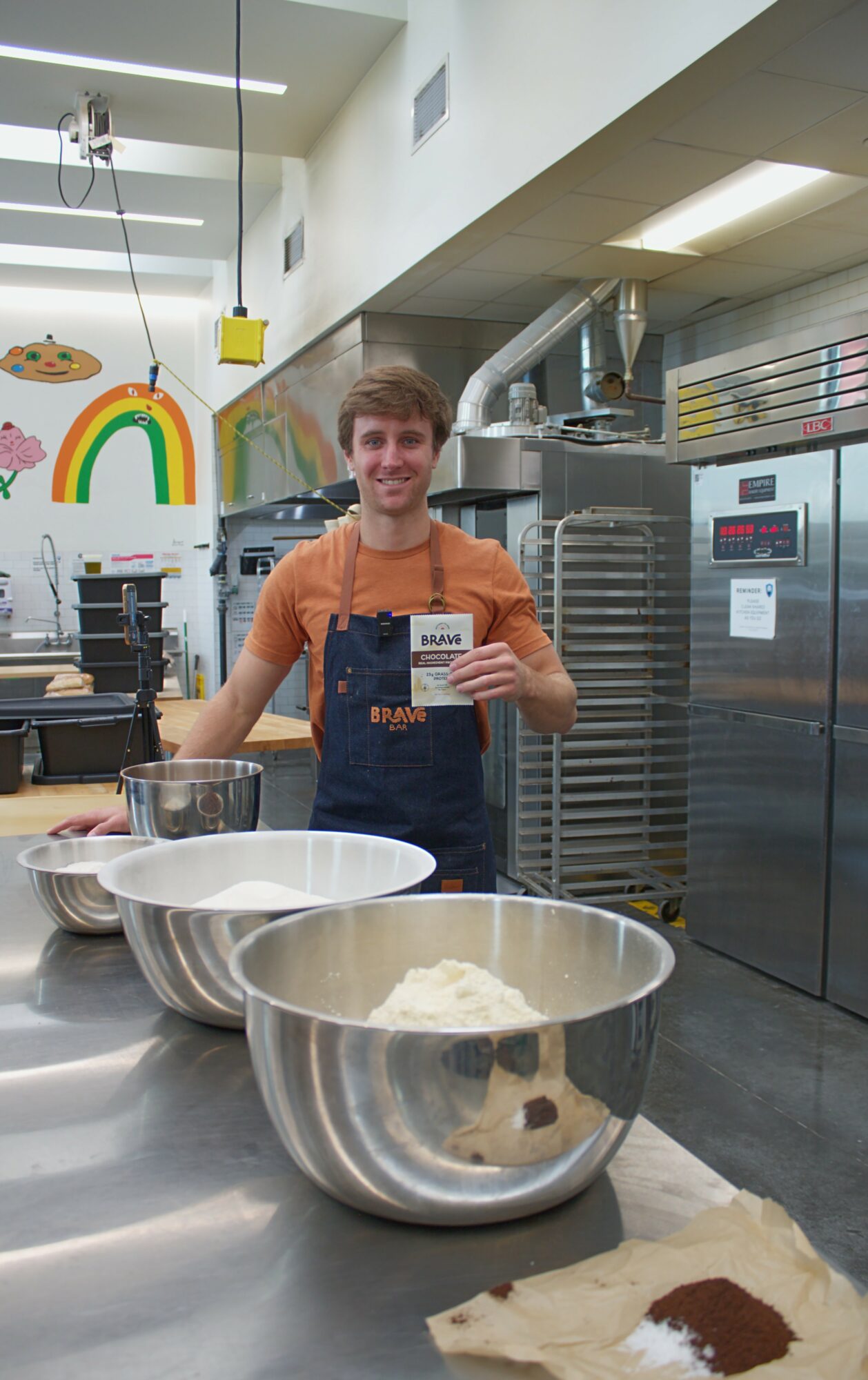 Young man in a kitchen holding a card, with large mixing bowls and baking ingredients on the counter.