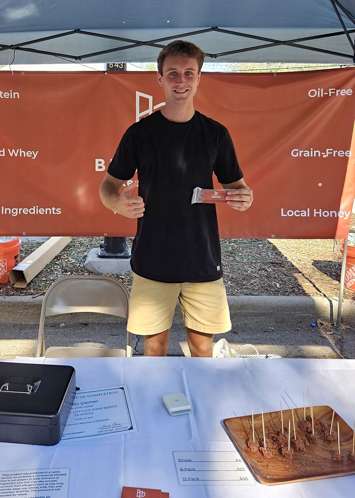 Young man standing behind a table at an outdoor booth, smiling, holding a small item, with a banner in the background.