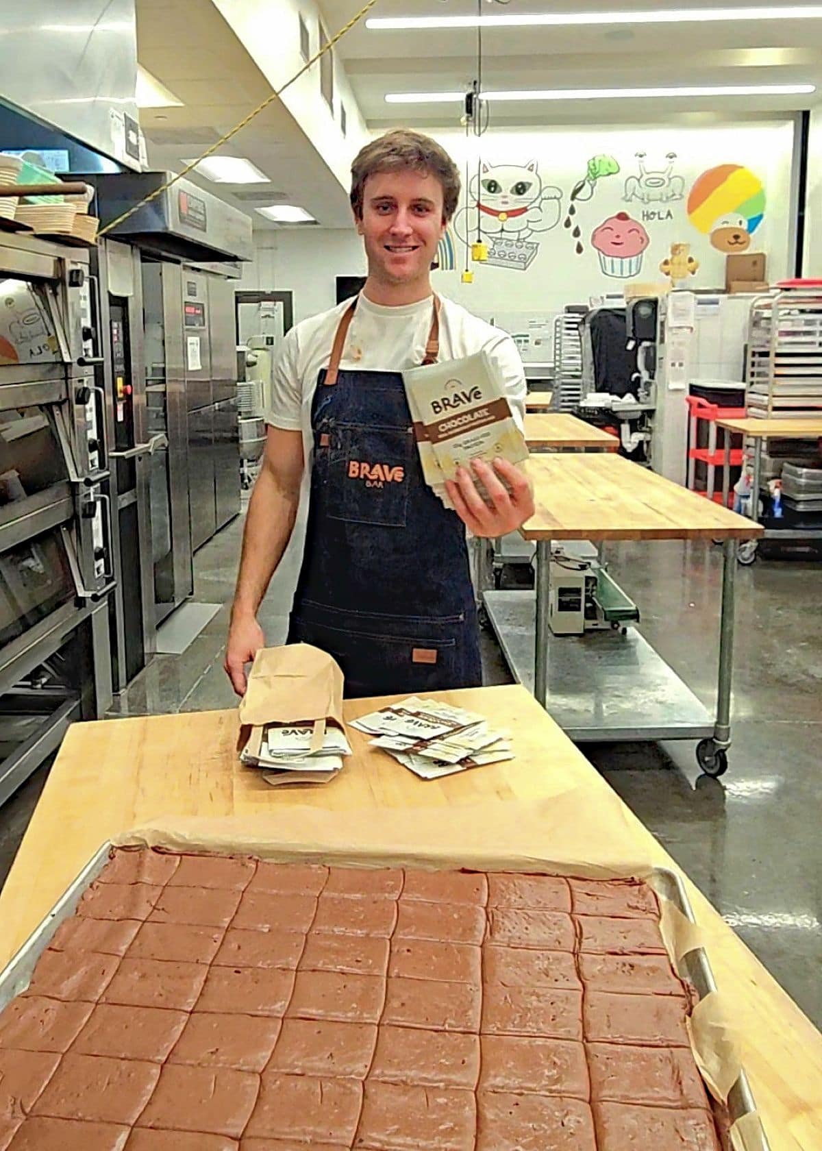 Young man in a kitchen holding a box of brownies, standing behind a table with baked brownies, smiling.