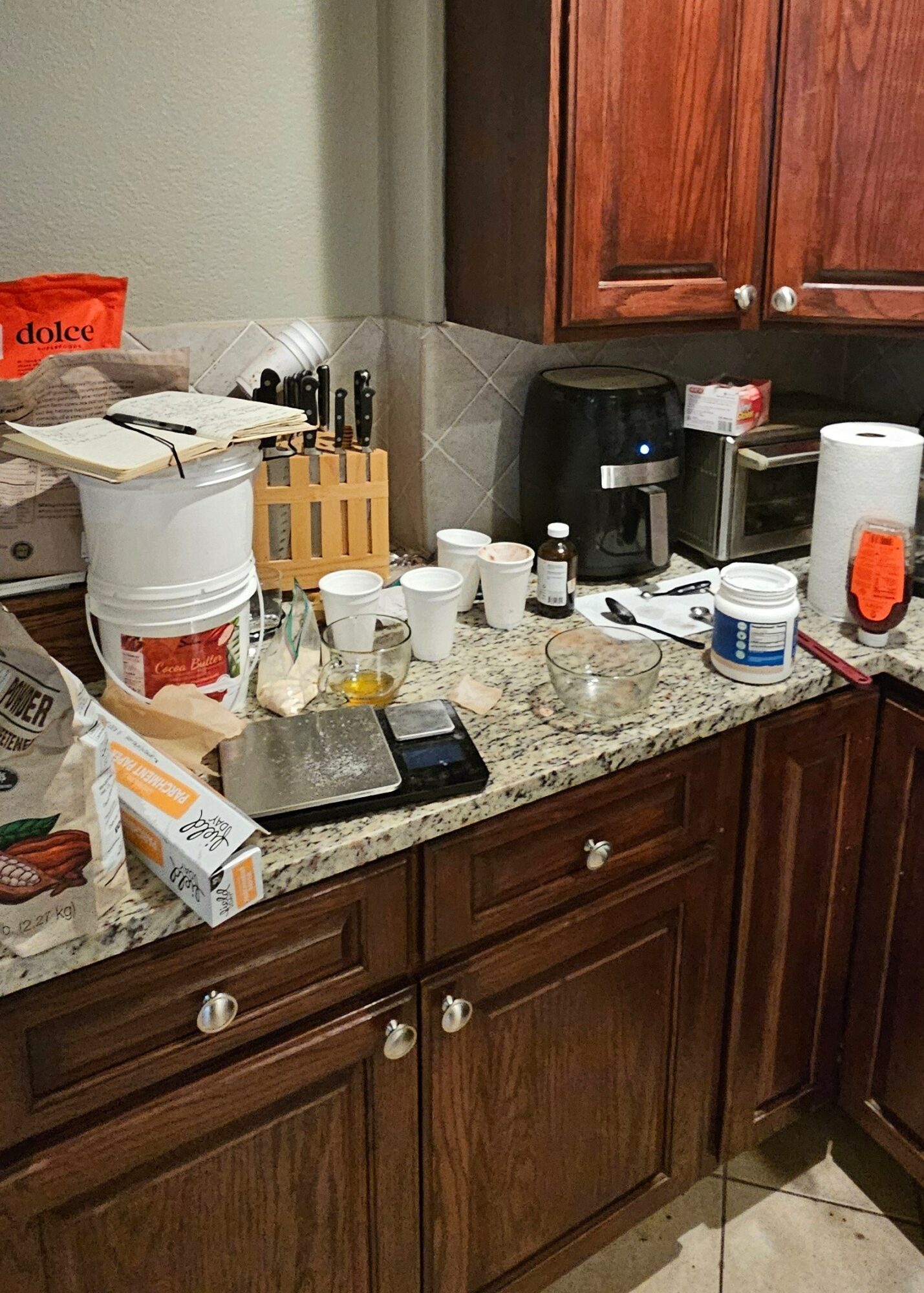 Kitchen countertop with coffee maker, cups, paper towels, and various items, with wooden cabinets above and below.