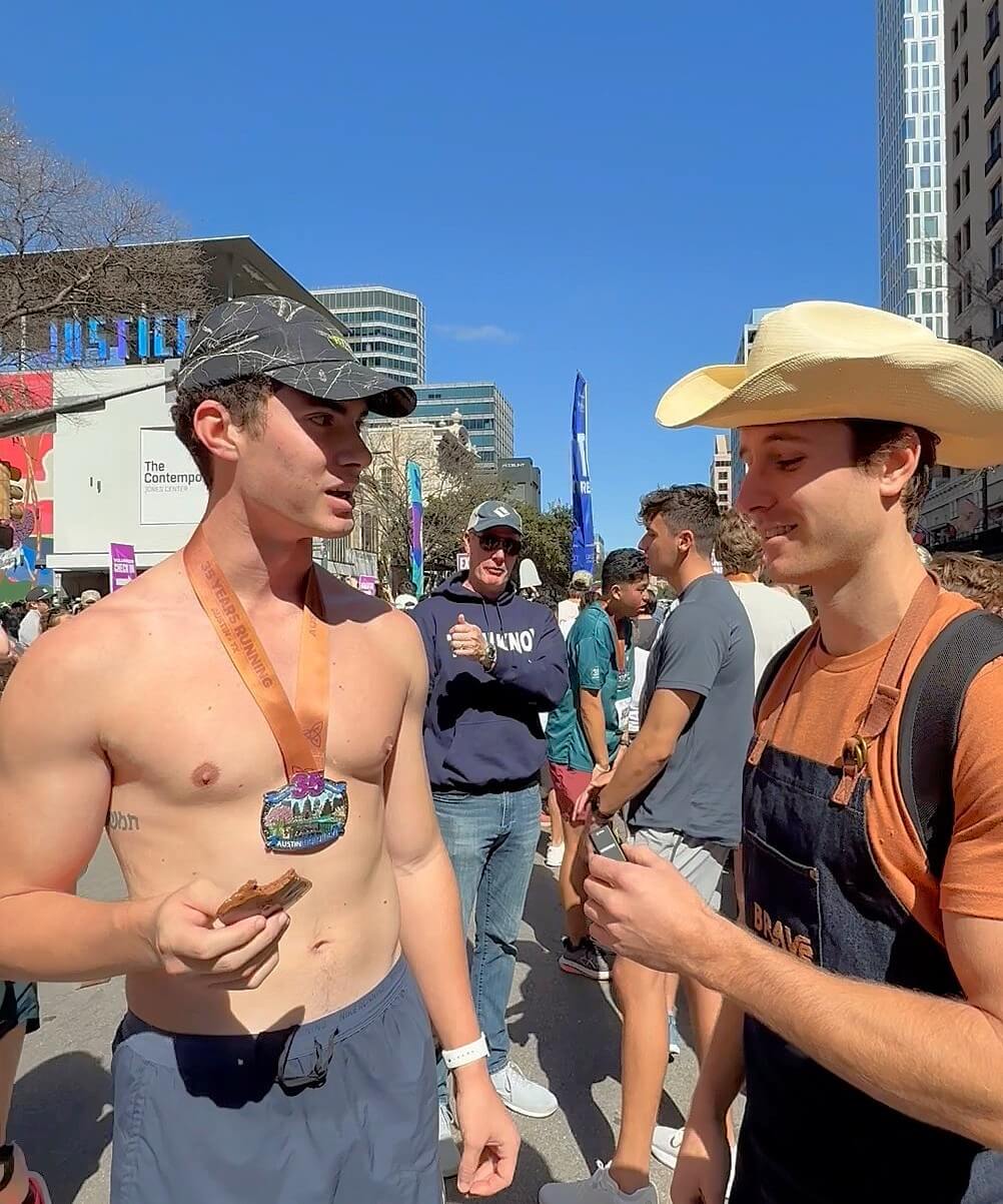Two young men with medals around their necks talking outdoors in a city square, surrounded by people and tall buildings.