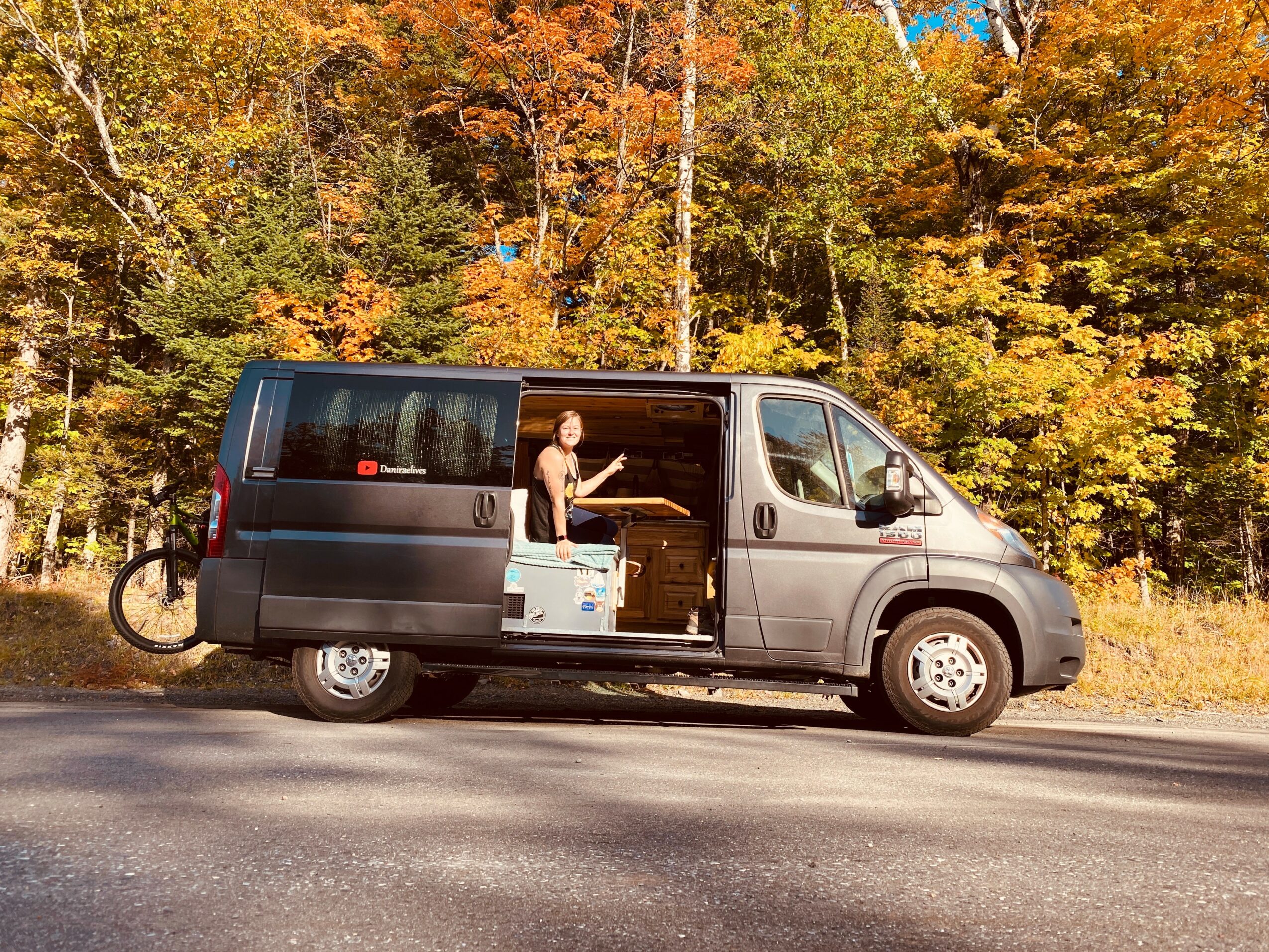 Person standing inside a black van with open sliding door, surrounded by autumn trees with colorful leaves.