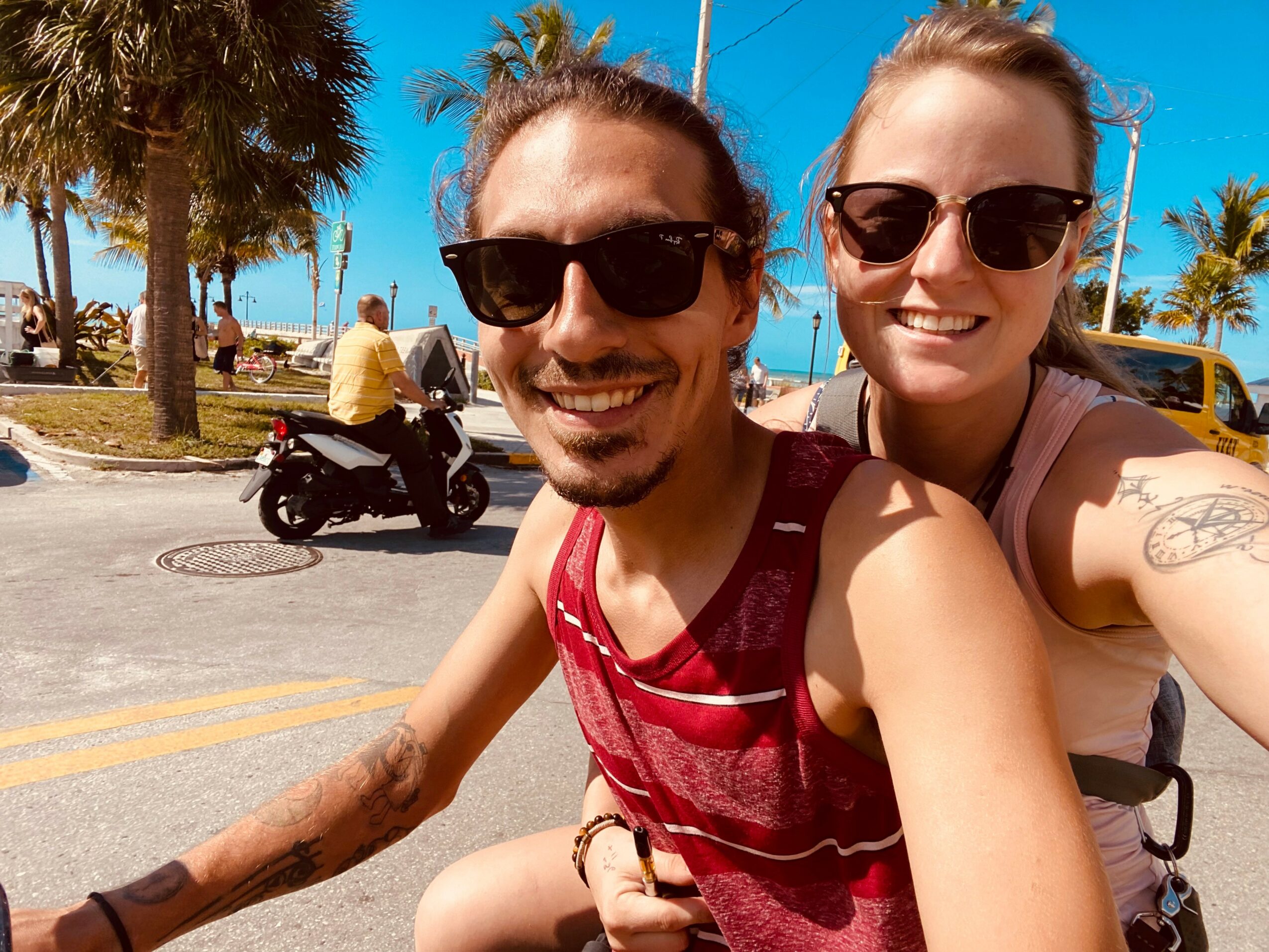 Two smiling people wearing sunglasses taking a selfie outdoors on a sunny day, with palm trees and a street in the background.
