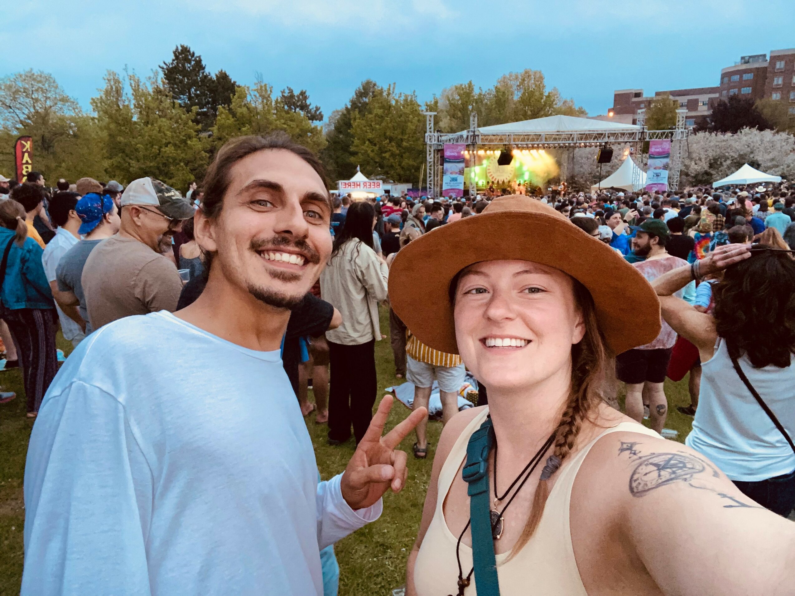 Two smiling people taking a selfie at an outdoor event with a crowd and stage in the background.