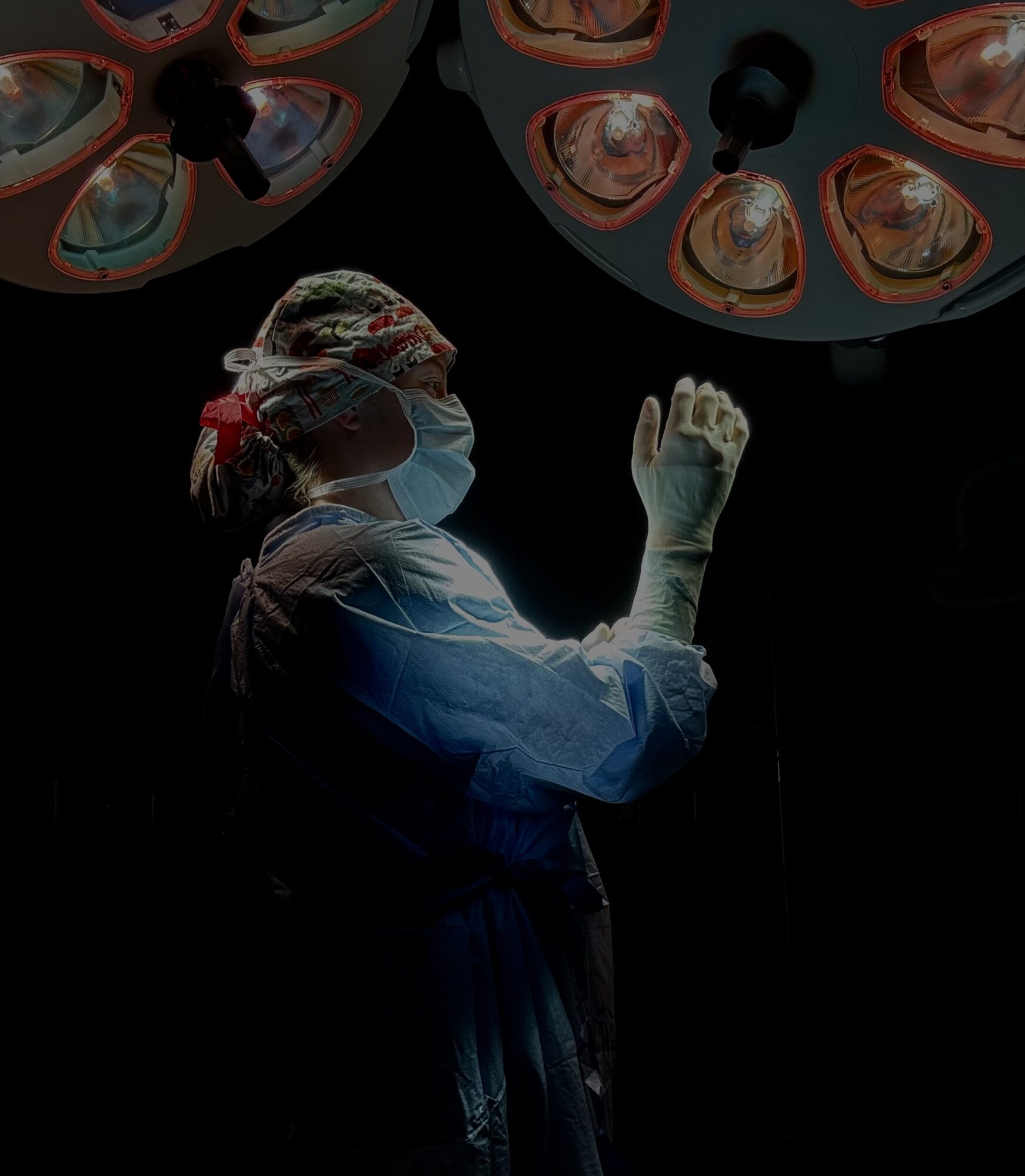 Surgeon in scrubs and mask raises hand under surgical lights in operating room.