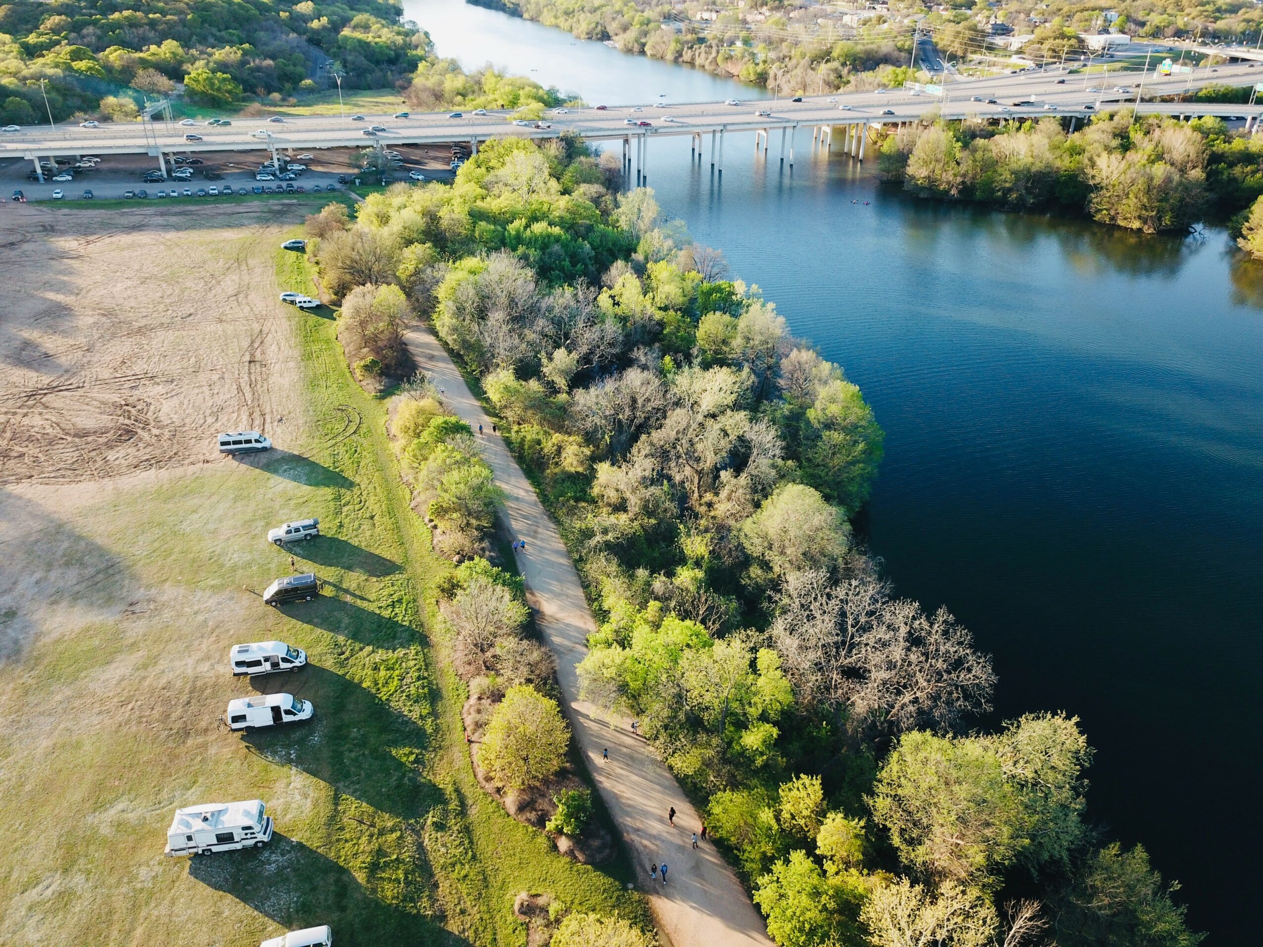 Aerial view of a river with trees along the shoreline and a park with parked cars and a walking path.