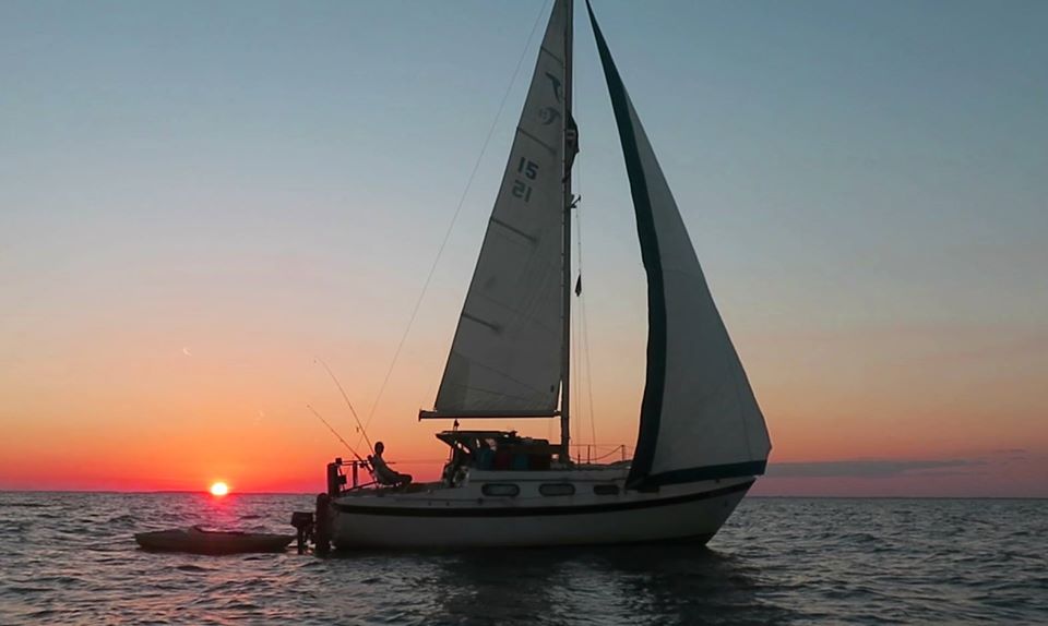 Sailboat on water during sunset with colorful sky and calm sea, silhouette of boat and crew visible.