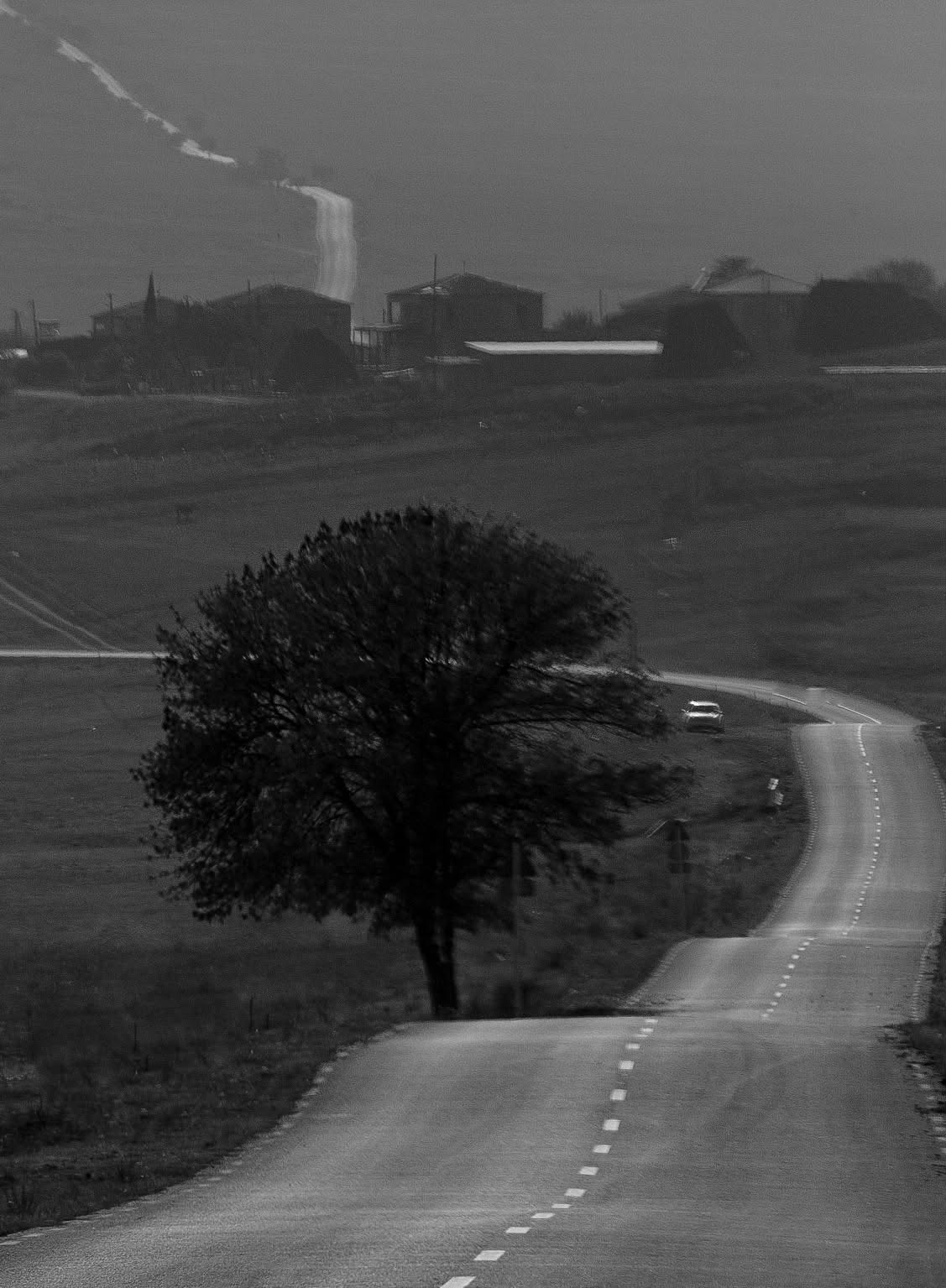 A winding road with a large tree in the foreground, rolling hills, and industrial buildings in the background, all in black and white.