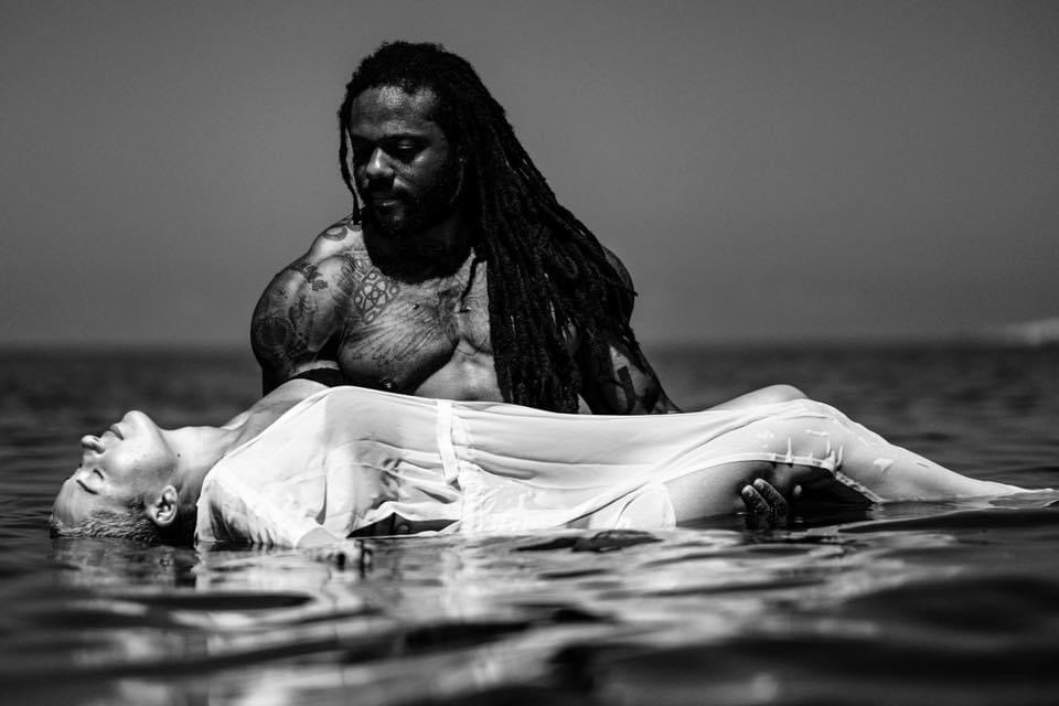Man with long dreadlocks holds a woman lying on water's surface, both in black and white, outdoors.