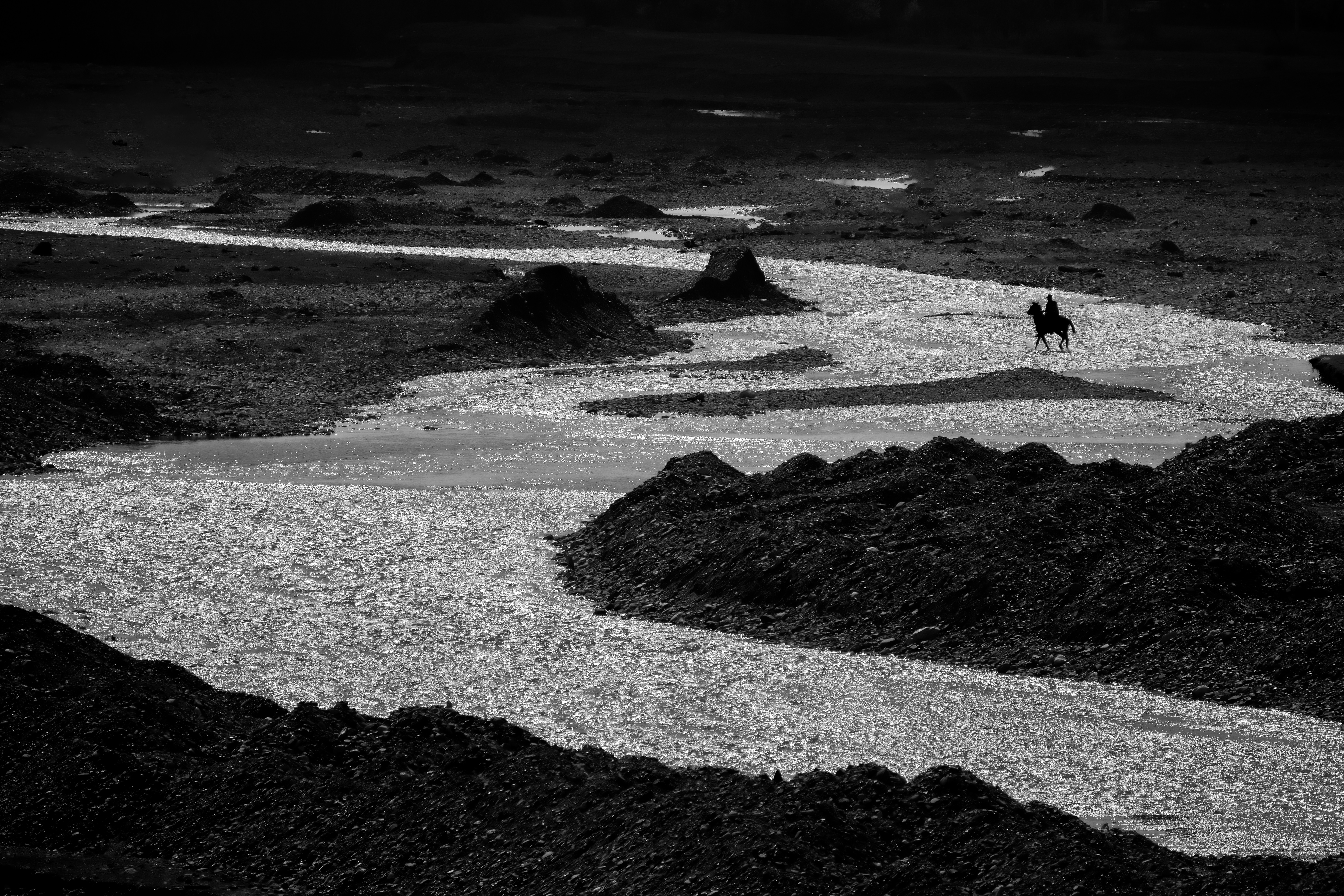 A black and white landscape with a winding river, rocks, and a distant horse, under a dark sky.