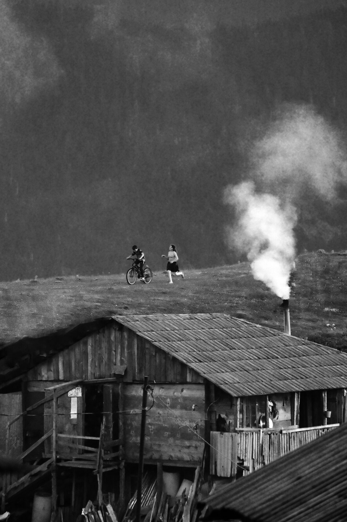 Two people with bicycles near a chimney emitting smoke, in front of a wooden building, landscape background.