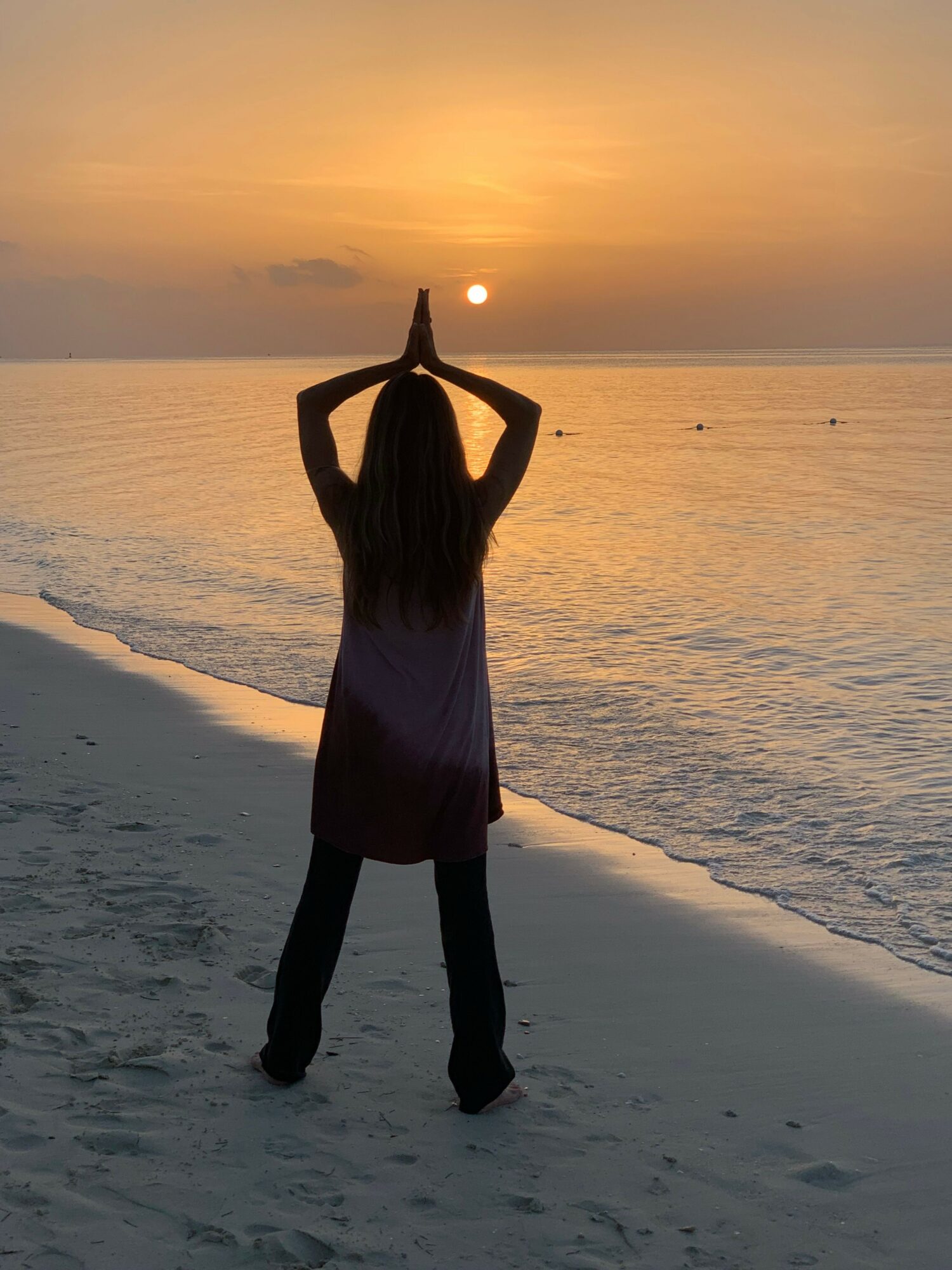 Person standing on beach at sunset, with hands forming a heart shape around the sun, facing the ocean.