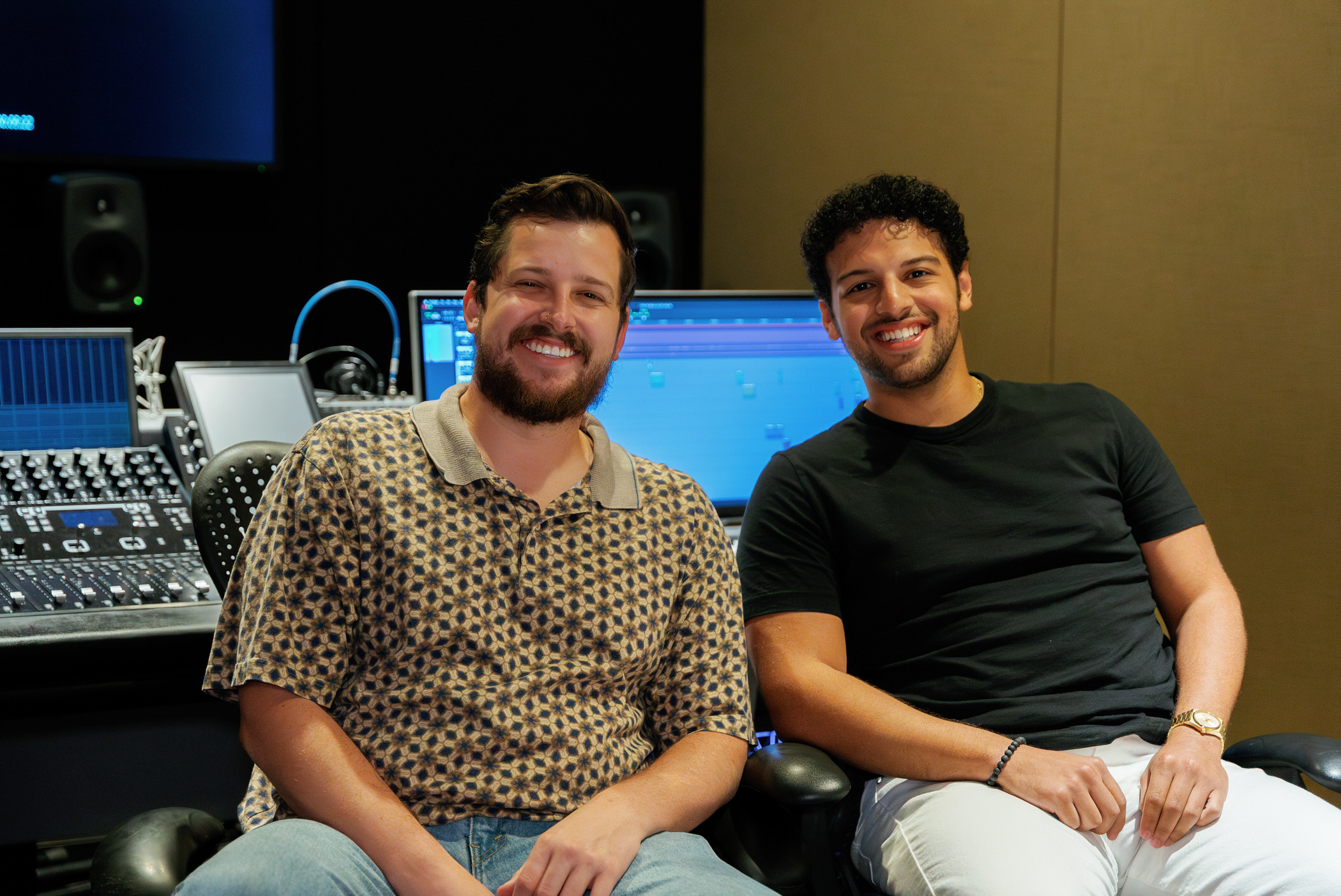 Two smiling men sitting in a recording studio with audio equipment and a computer screen behind them.