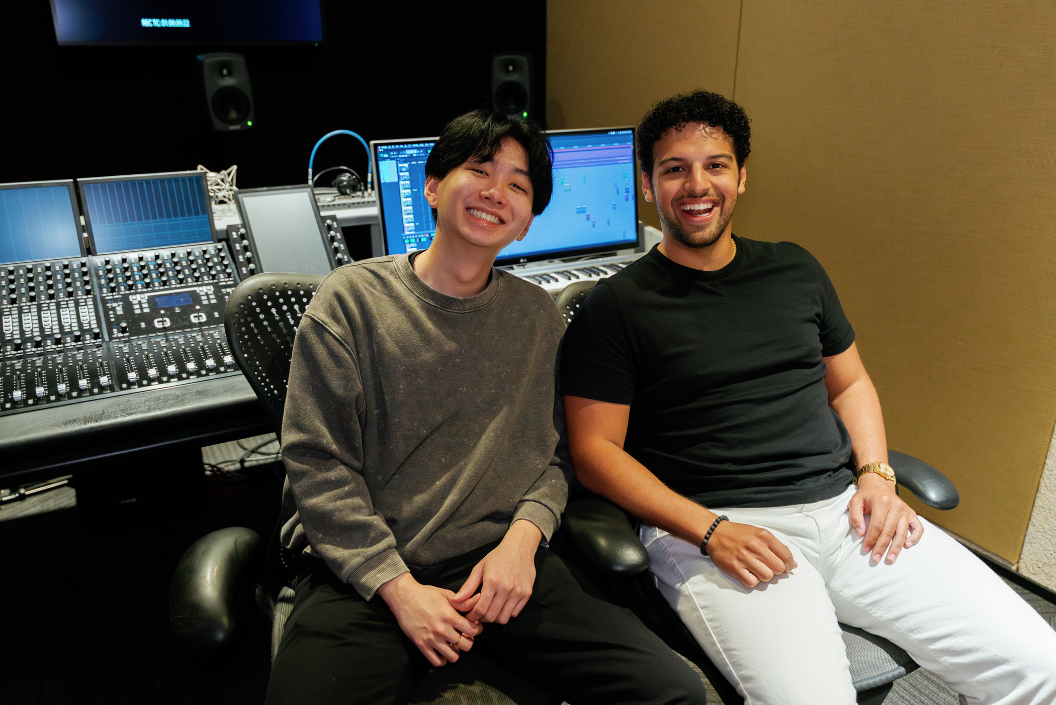 Two people sitting in a recording studio with audio equipment and computer screens behind them.