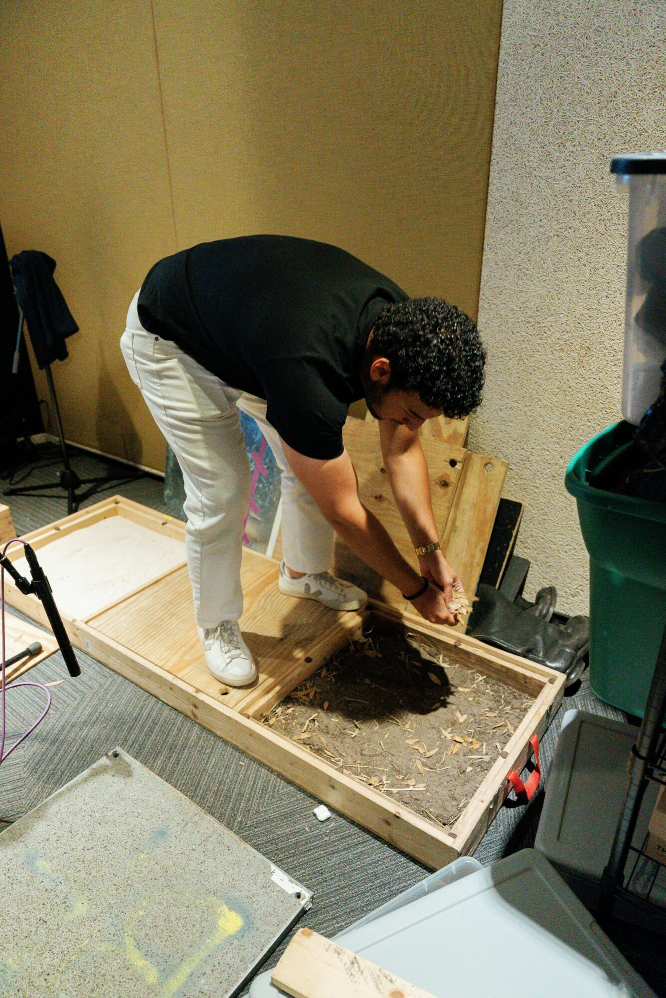 Person working on a gardening project, planting soil in a wooden frame, indoors with various tools and containers nearby.