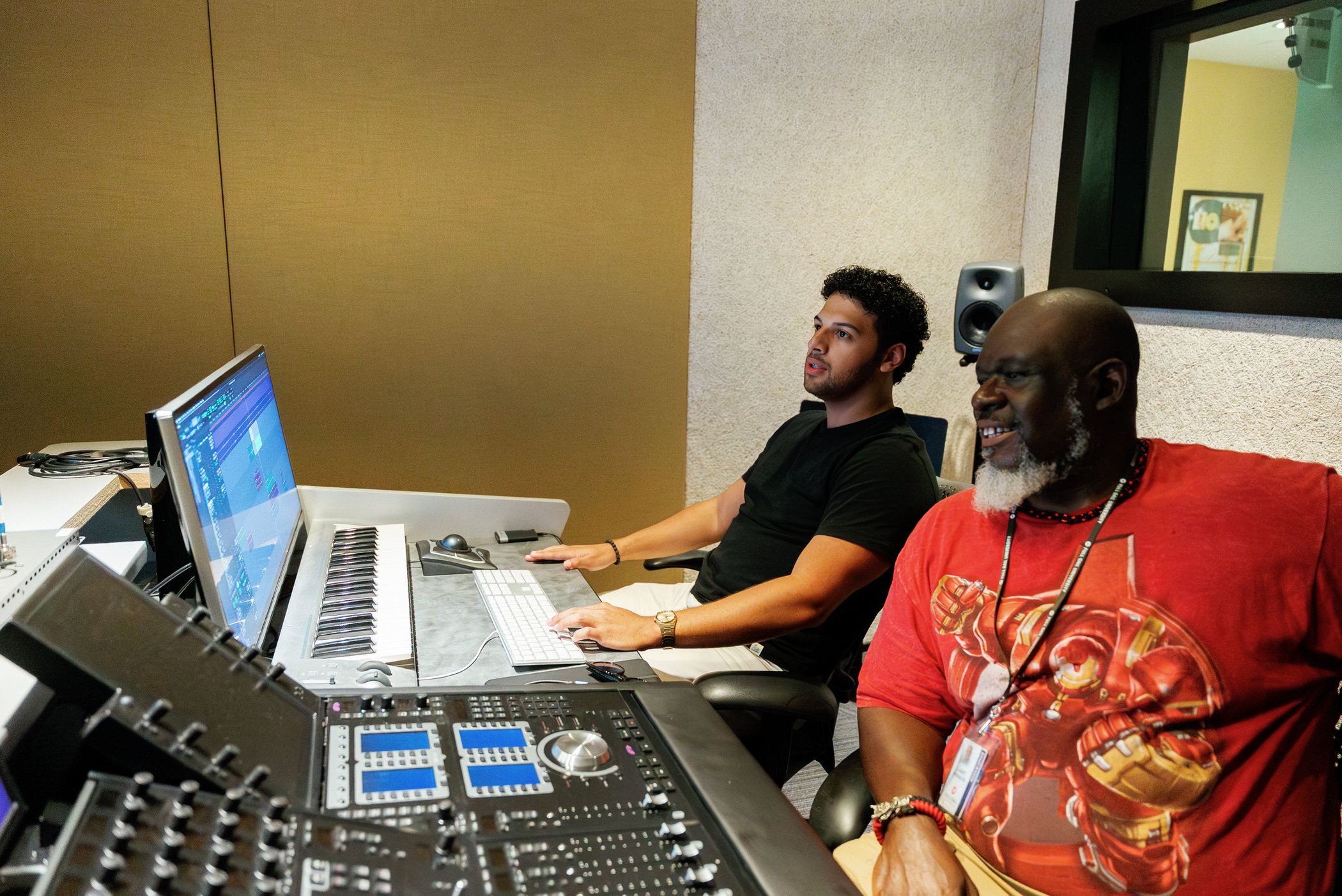Two men sit in a recording studio, one at a computer and the other at a mixing console, with studio equipment visible.