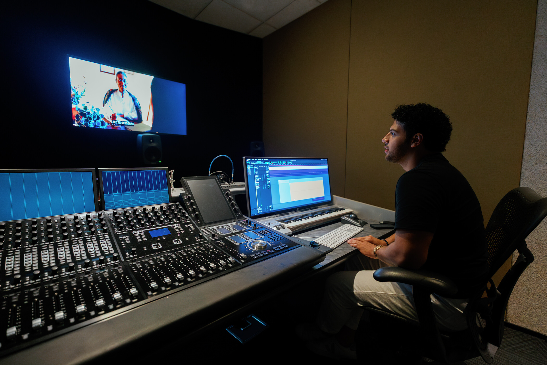 Person working at audio mixing console in a recording studio, looking at a computer screen, with a monitor displaying a colorful image.