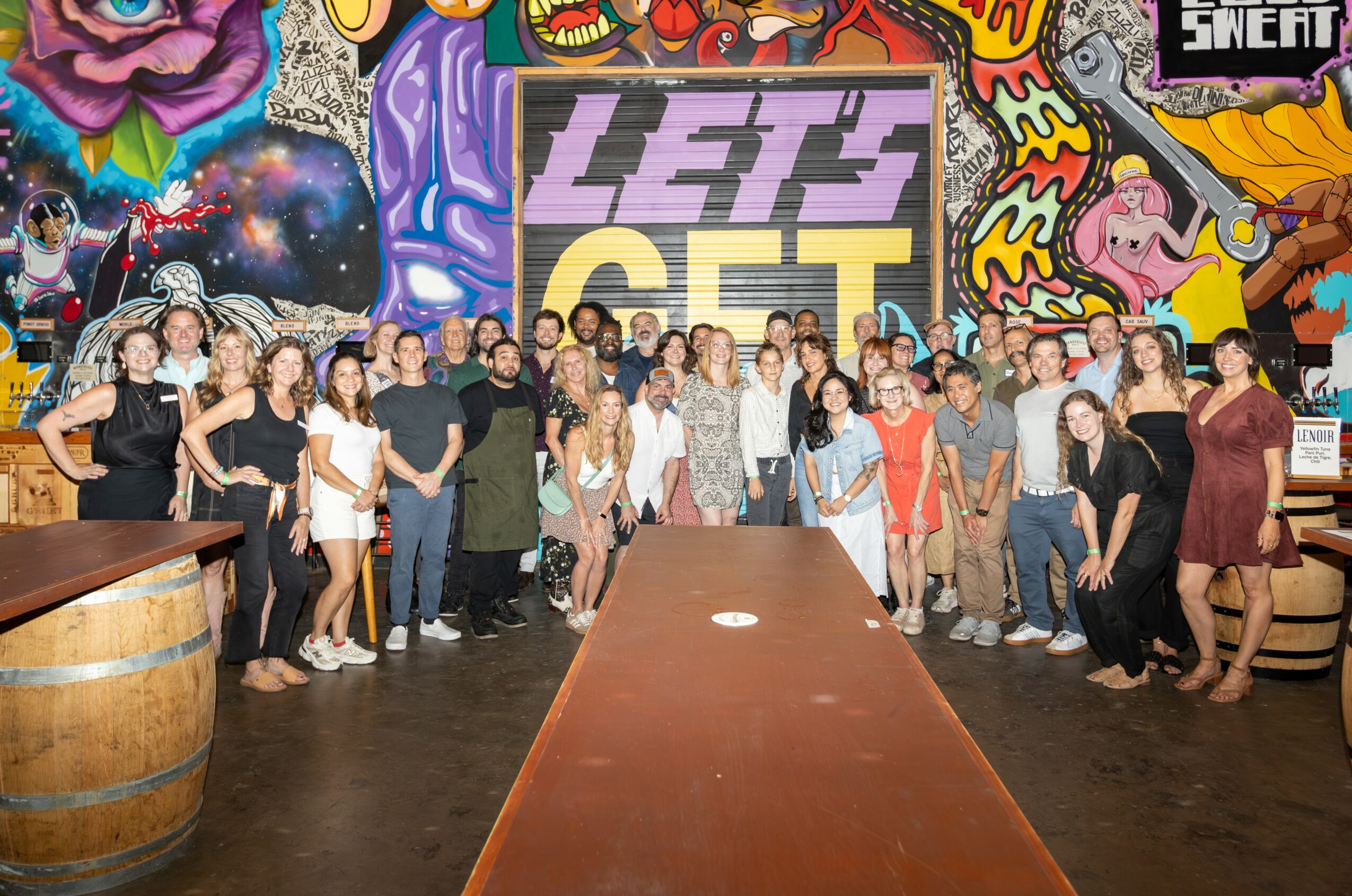 Group of people standing in front of colorful graffiti wall, some smiling, some with hands in pockets, in a room with a long table.
