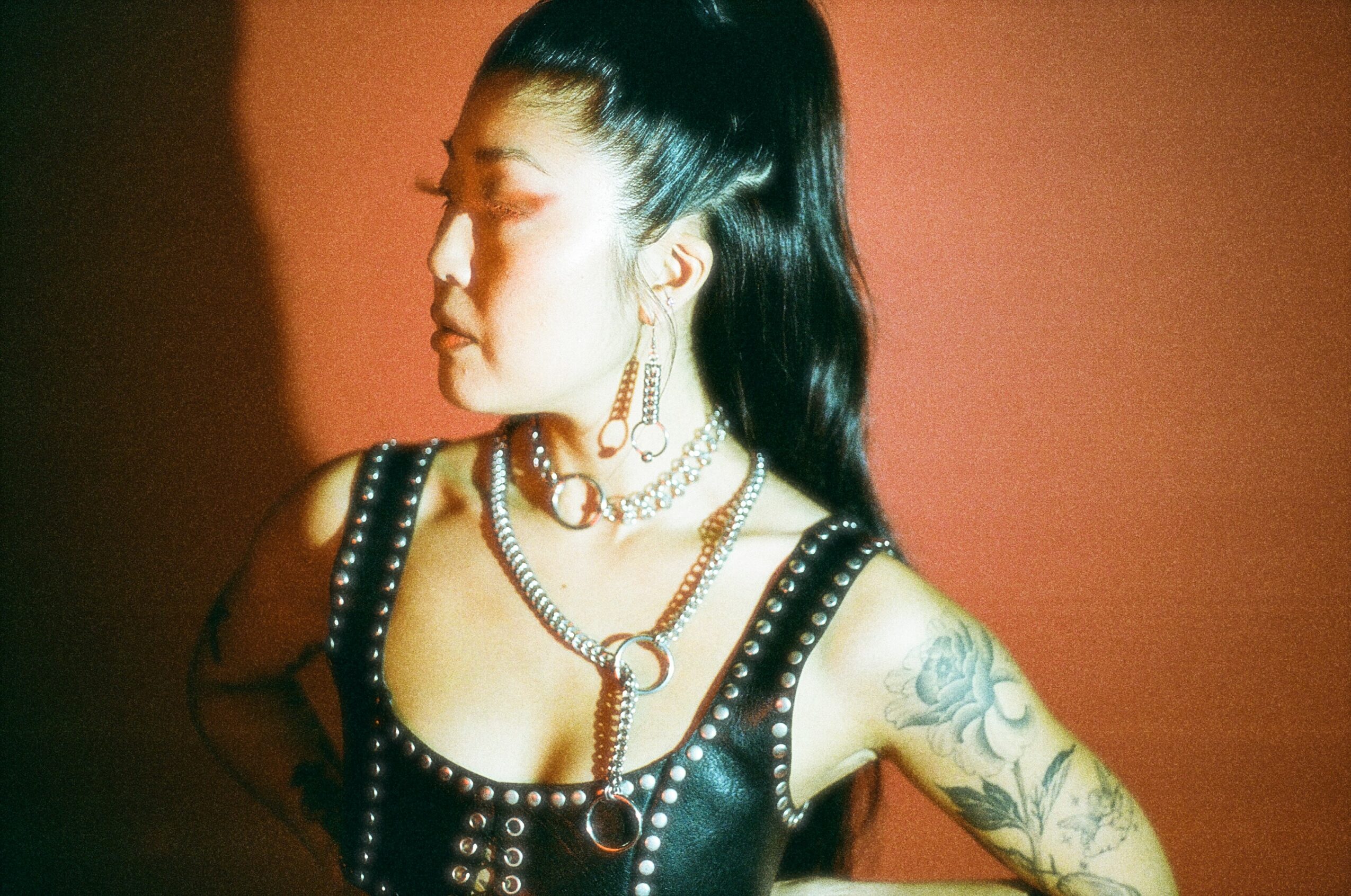 Woman with long dark hair in a leather outfit with jewelry, looking to the side against a reddish background.