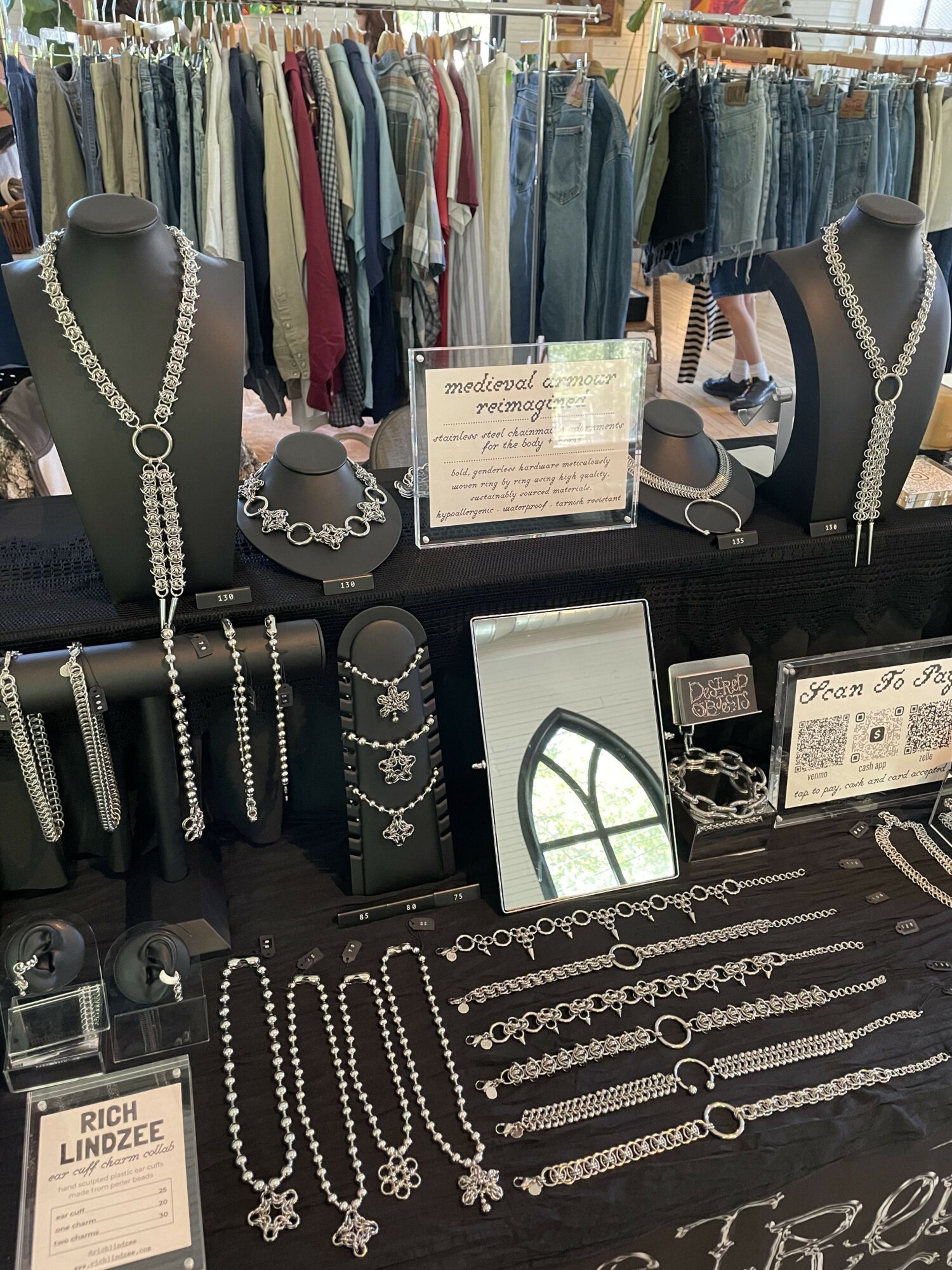 Jewelry display with necklaces, bracelets, and earrings, set against a clothing rack with jeans and shirts in the background.