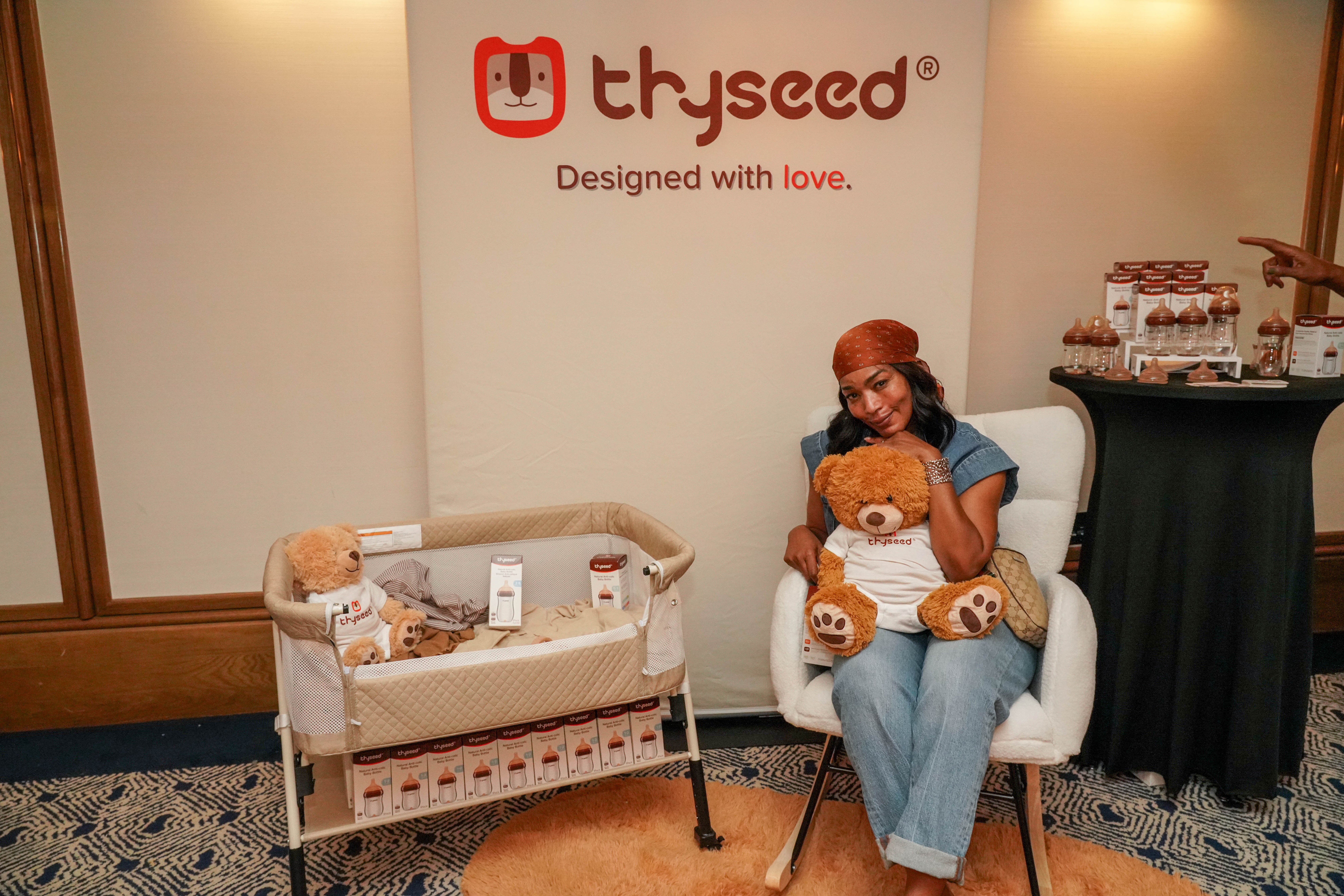 Woman sitting on a white chair holding a teddy bear, with a baby crib nearby and a display table in the background.